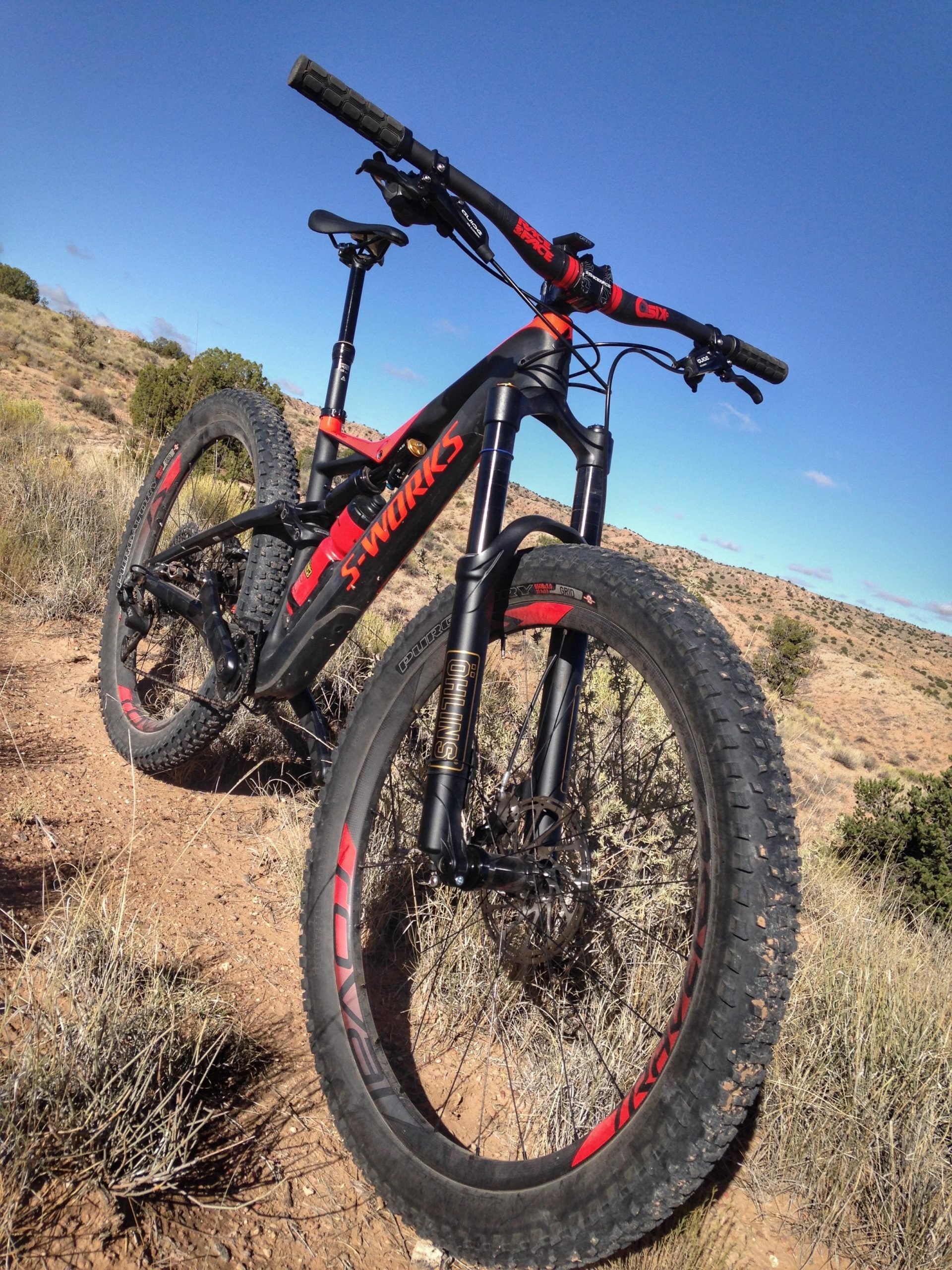 A mountain bike with a sleek black and red design stands on a dirt trail surrounded by sparse vegetation and hills under a clear blue sky. The bike features large, knobby tires and a high-performance suspension system, emphasizing its suitability for rugged terrain. Mariposa Fat Bike Trails mountain bike trail.