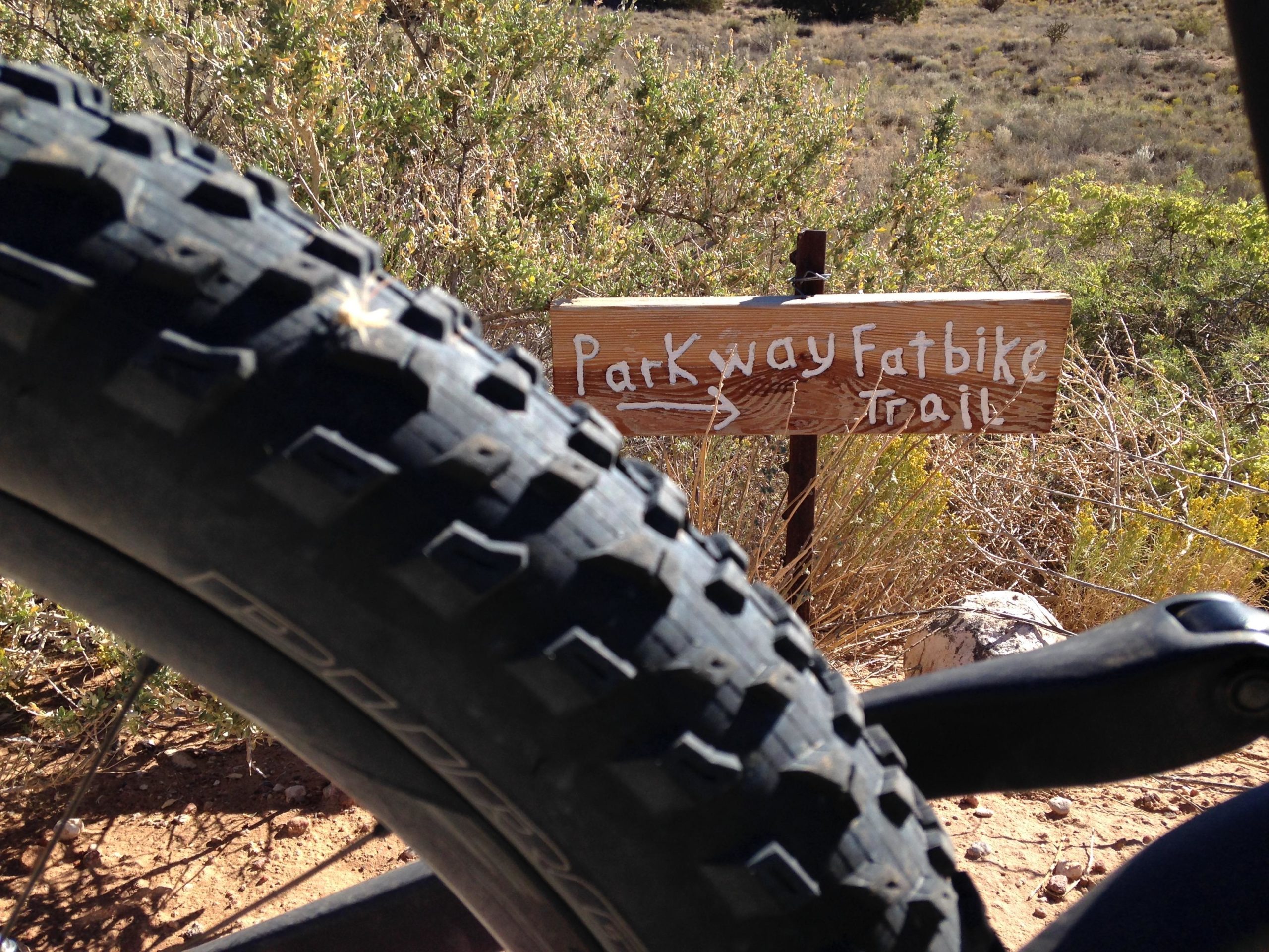 A close-up view of a fat bike tire in the foreground with a wooden trail sign in the background that reads "Parkway Fatbike Trail." The scene is set in a natural landscape with shrubs and dry terrain visible. Mariposa Fat Bike Trails mountain bike trail.