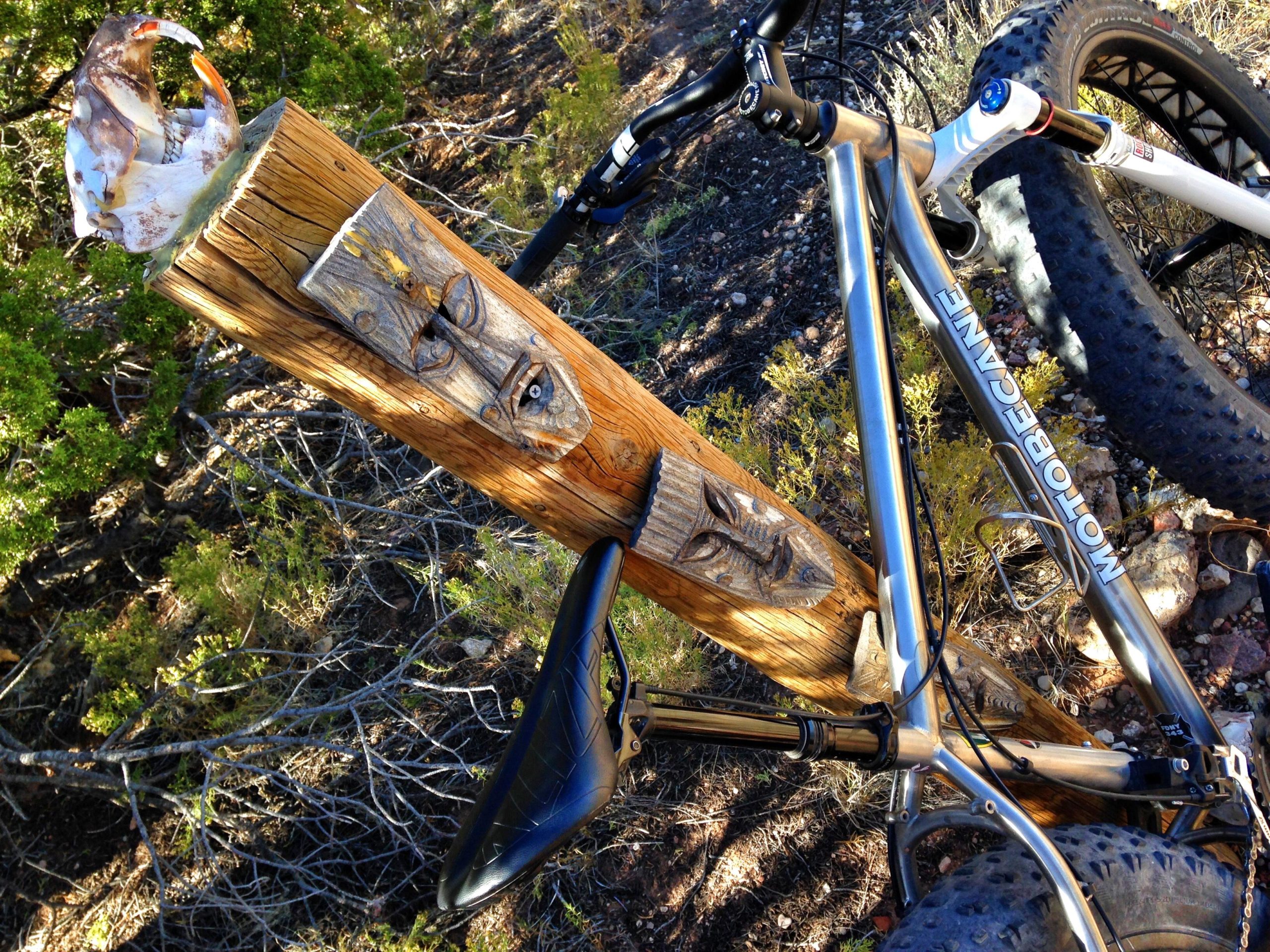 A mountain bike resting against a carved wooden post in a natural outdoor setting, featuring a decorative shell on top and intricate carvings resembling faces. Surrounding vegetation includes shrubs and small plants. Mariposa Fat Bike Trails mountain bike trail.