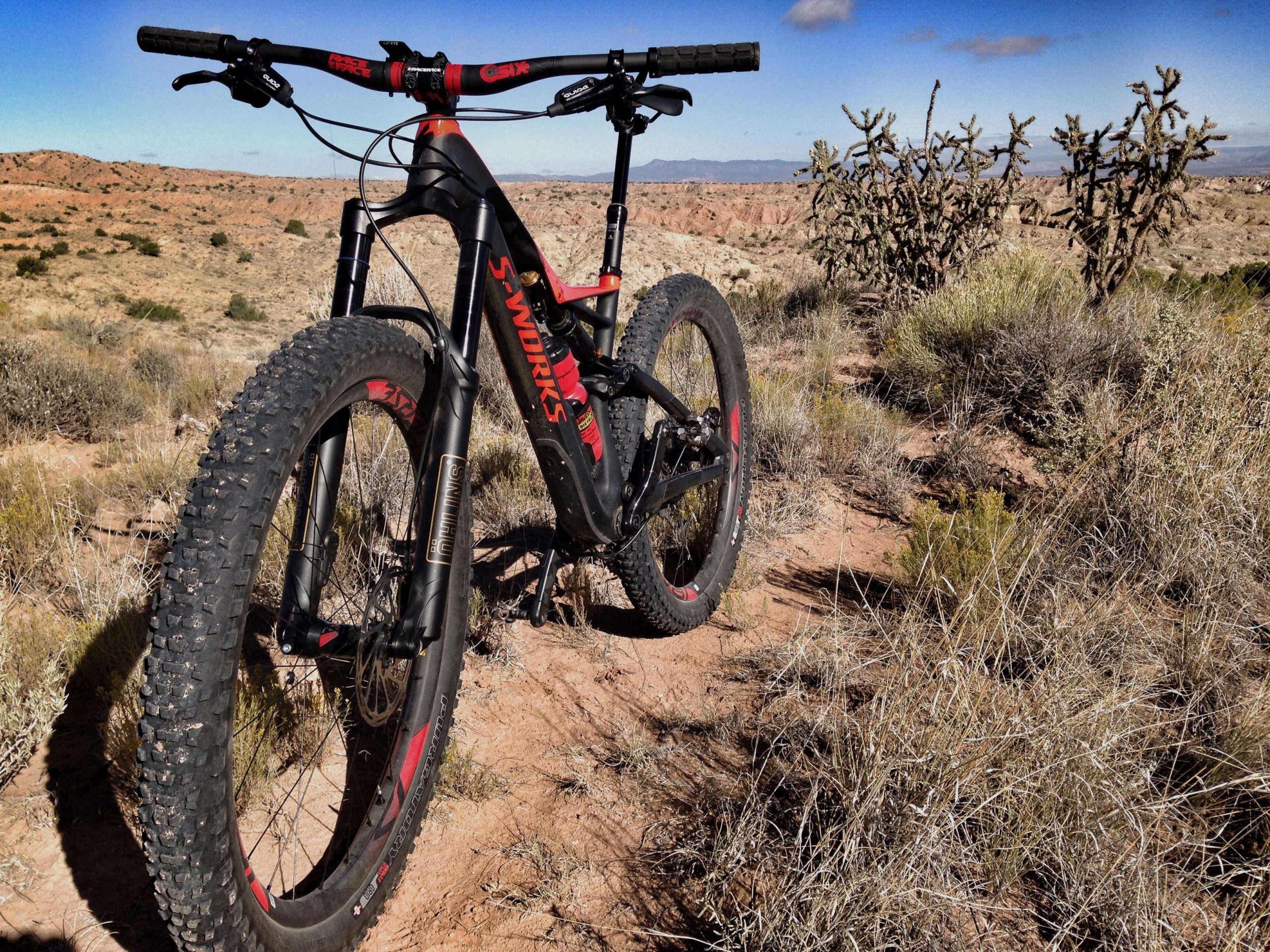 A mountain bike with bold red accents stands on a dirt trail, surrounded by rugged terrain and sparse vegetation. In the background, a rocky landscape stretches out under a bright blue sky with a few clouds. The bike's front tire is prominent in the foreground, showcasing its sturdy design, while the natural scenery emphasizes the outdoor adventure setting. Mariposa Fat Bike Trails mountain bike trail.