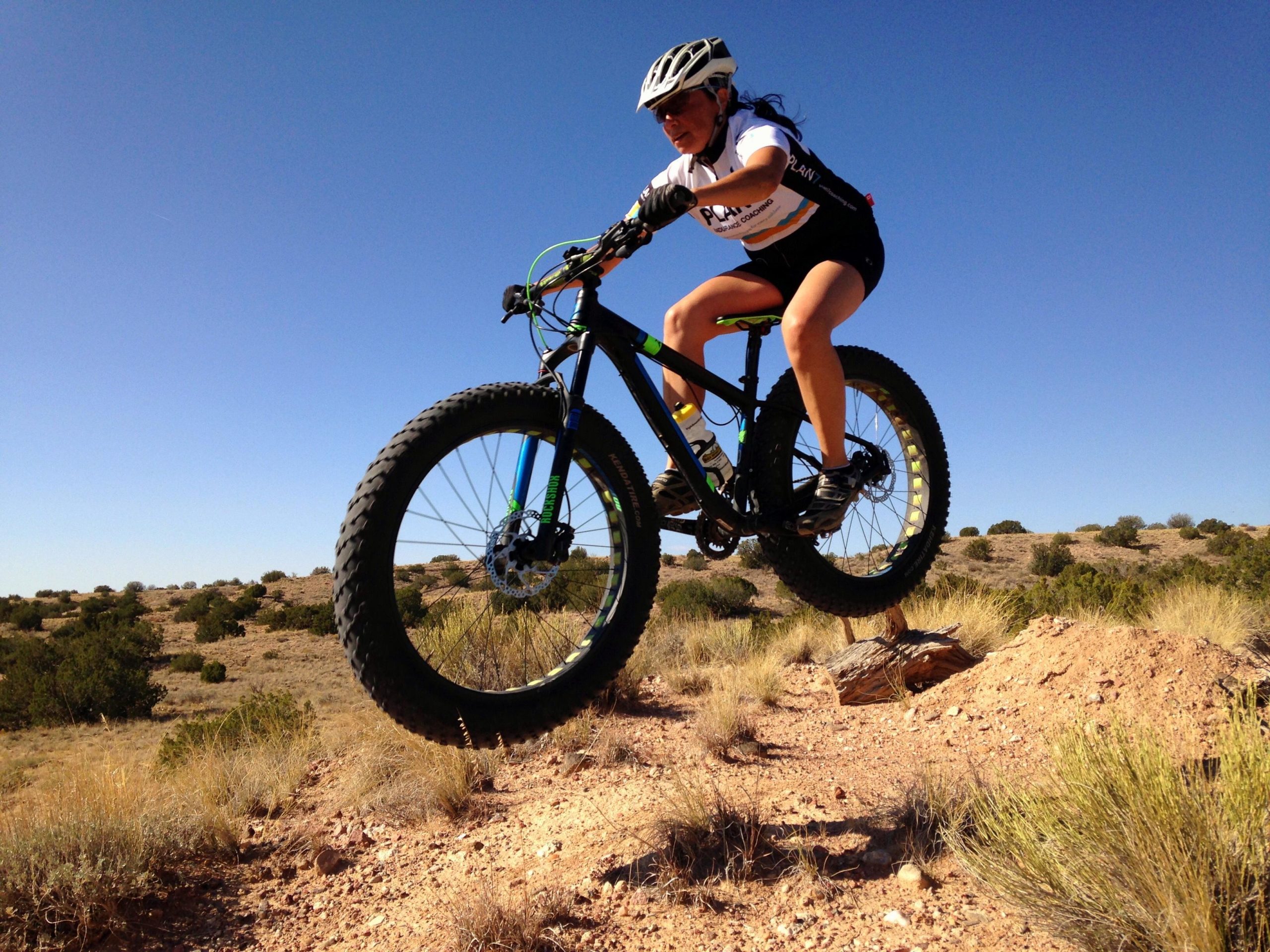 A mountain biker in mid-air jumps over a rocky terrain, showcasing a dynamic action shot. The rider wears a helmet and a cycling jersey, and is riding a fat bike on a sunny day with a clear blue sky in the background. Surrounding the scene are dry shrubs and rolling hills. Mariposa Fat Bike Trails mountain bike trail.
