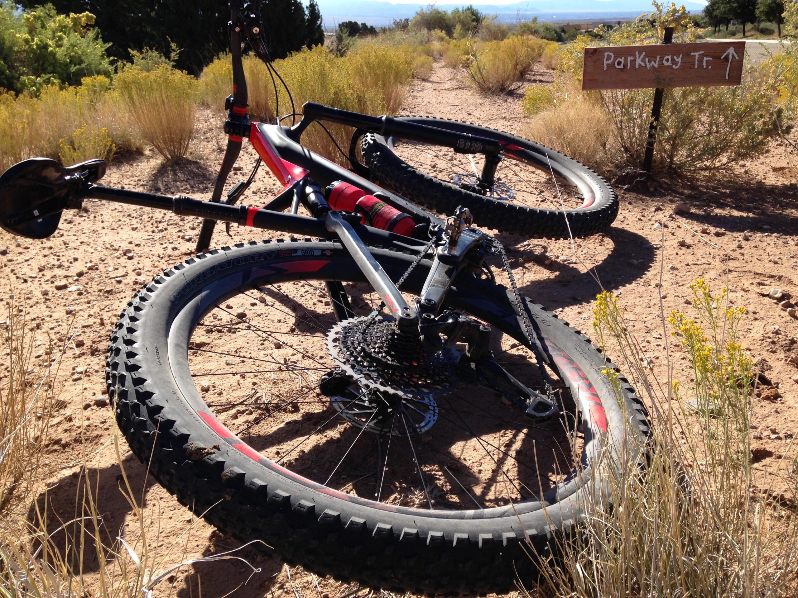 A mountain bike lies on its side on a dirt trail surrounded by tall grass and shrubs. In the background, a wooden sign points towards "Parkway Trail," indicating the direction for hikers or bikers. The scene is set in a sunny, outdoor environment. Parkway Fatbike trail mountain bike trail.