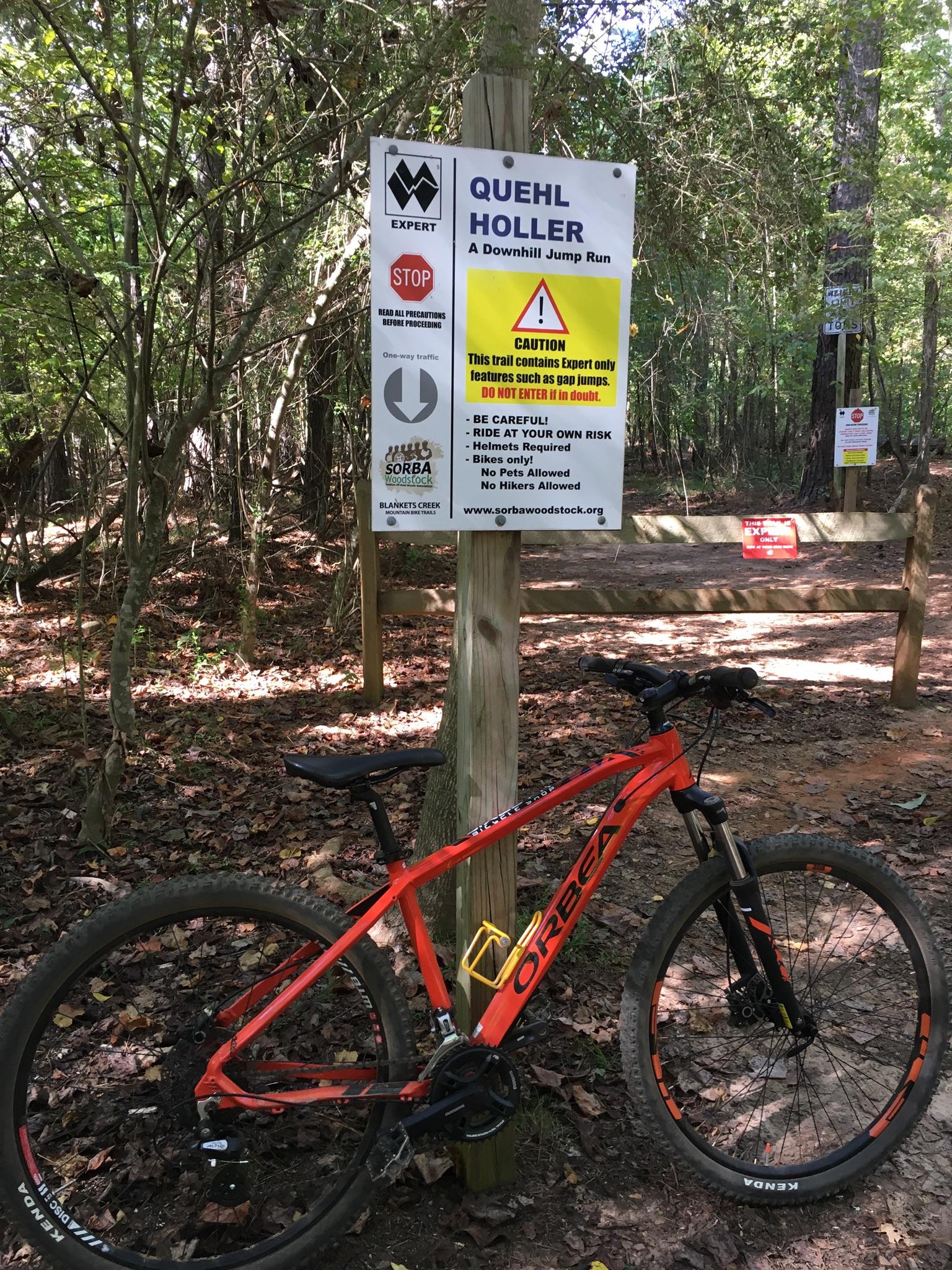 A mountain bike leans against a wooden post, next to a sign indicating the start of "Quehl Holler," an expert-level downhill jump run. The sign features warnings about the trail's risks, including that it contains gap jumps, and advises riders to wear helmets and ride at their own risk. The surrounding area is wooded with fallen leaves on the ground. Blankets Creek mountain bike trail.