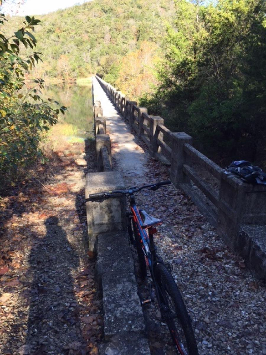 A mountain bike is placed on a stone path beside a narrow bridge that extends over a calm body of water, surrounded by trees in autumn foliage. A shadow of a person is visible on the gravel surface, indicating presence nearby. The scenery features hills in the background, adding to the natural setting. Lake Leatherwood Gravity Project mountain bike trail.
