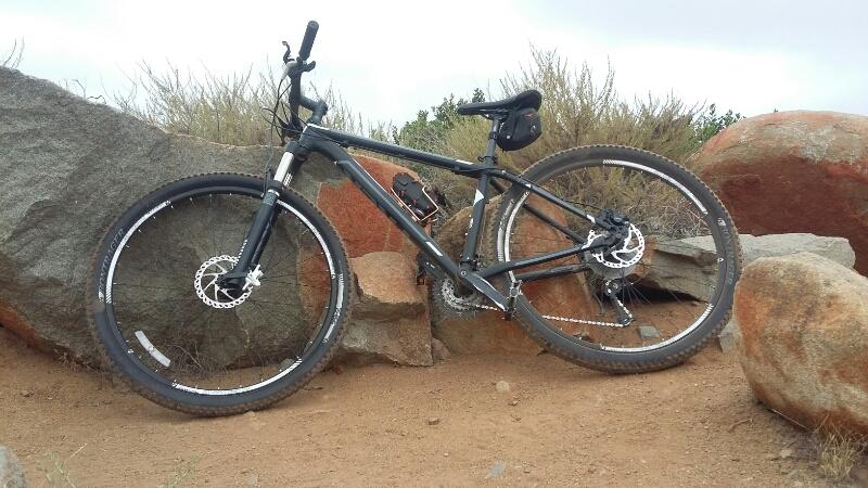 Trek Mamba: A black mountain bike leaning against large rocks in a sandy outdoor setting, with sparse vegetation in the background under a cloudy sky.