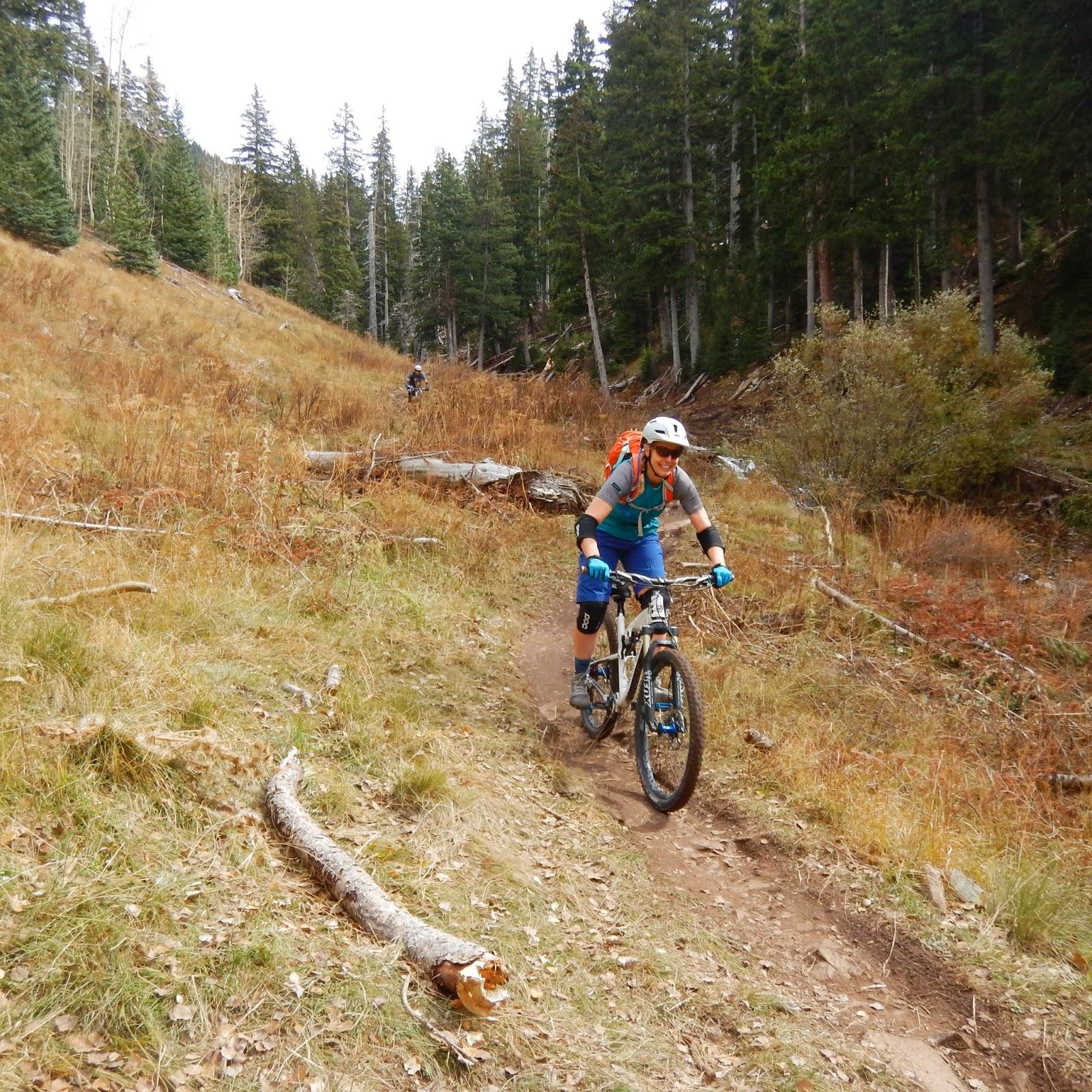A person riding a mountain bike on a dirt path surrounded by tall trees and autumn foliage. The cyclist is wearing a helmet, gloves, and a colored shirt and shorts, smiling as they navigate the trail. In the background, another cyclist is visible. The Whole Enchilada mountain bike trail.