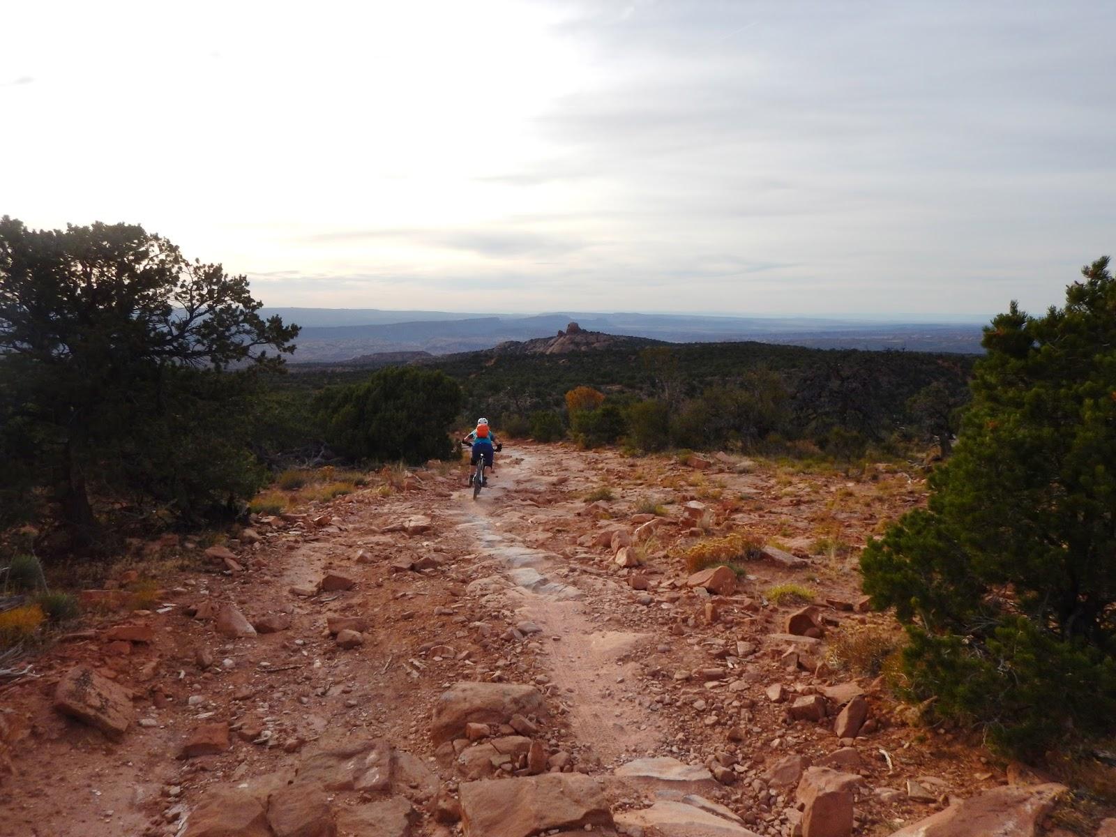 A person riding a mountain bike on a rocky trail surrounded by sparse vegetation and trees, with a scenic view of distant mountains under a cloudy sky. The Whole Enchilada mountain bike trail.