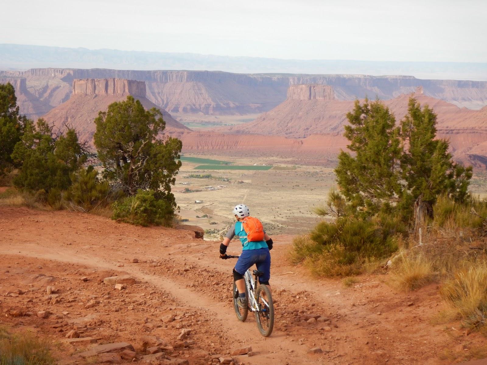 A mountain biker riding along a dirt trail with a scenic view of red rock formations and valleys in the background. The landscape features green fields and distant mesas under a clear sky. The Whole Enchilada mountain bike trail.