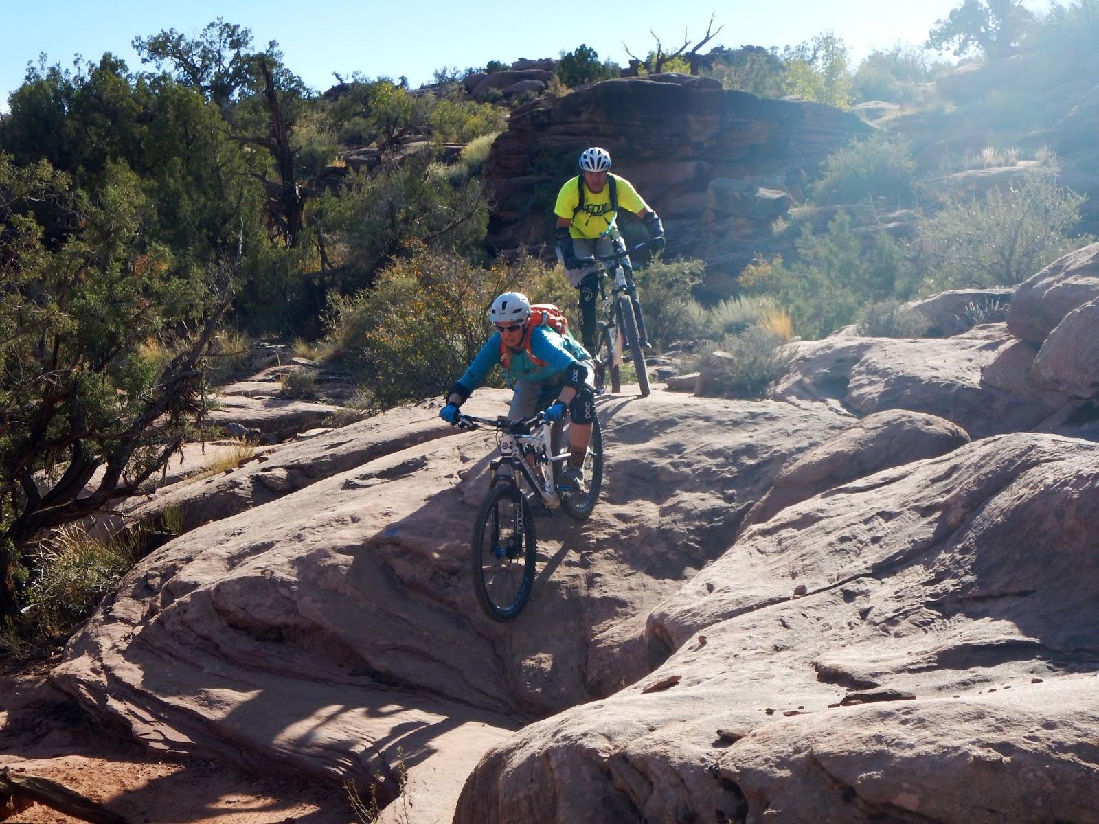 Two mountain bikers navigating a rocky trail in a sunlit outdoor setting, surrounded by trees and rugged terrain. The first rider, wearing a helmet and blue jersey, is approaching a rocky outcrop, while the second rider in a yellow shirt follows behind. Captain Ahab mountain bike trail.