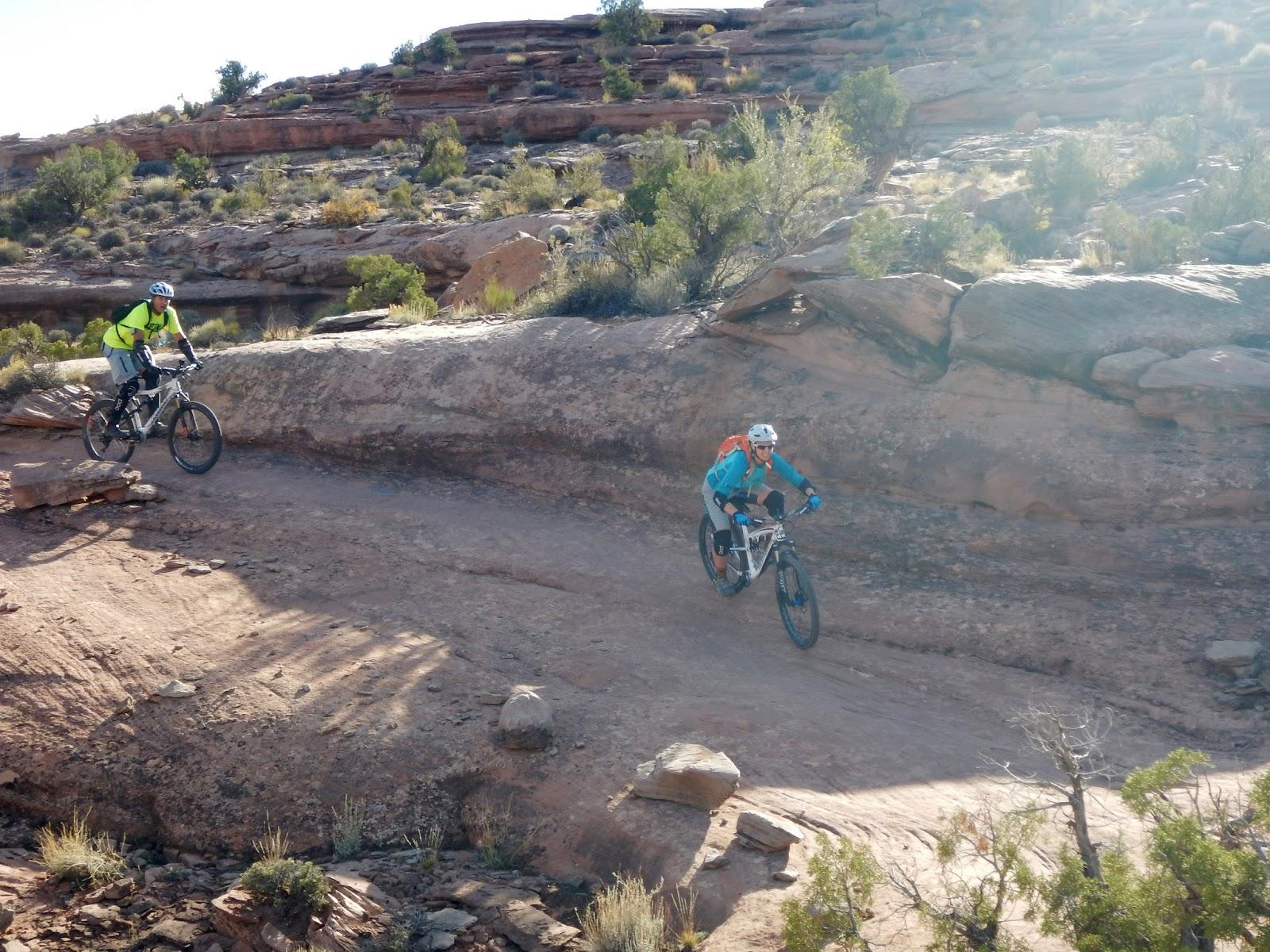 Two mountain bikers navigate a rocky trail in a desert landscape, surrounded by rugged terrain and sparse vegetation under a bright sky. One biker wears a bright yellow shirt and rides to the left, while the other, dressed in blue with an orange backpack, rides on the right side of the trail. Captain Ahab mountain bike trail.