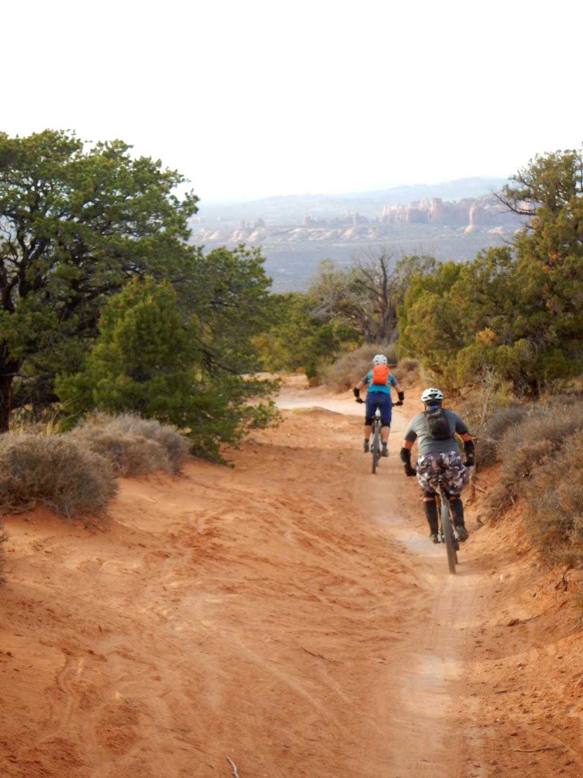 Two mountain bikers riding on a sandy dirt trail surrounded by vegetation, with a scenic view of distant rock formations and hills in the background. The Whole Enchilada mountain bike trail.