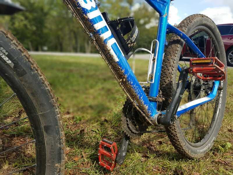 A close-up view of a blue mountain bike showing the frame, pedals, and muddy tires, positioned on a grassy area with trees and a road in the background. The bike frame has visible dirt splatters, indicating recent use on trails. Veterans Park mountain bike trail.