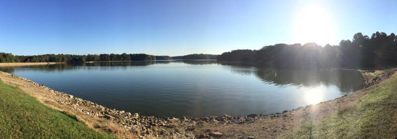 A panoramic view of a serene lake reflecting the clear blue sky, bordered by green grass and rocky shores. Sunlight glimmers on the water's surface, and a forested area is visible in the background, creating a peaceful natural scene. Herb Parsons mountain bike trail.
