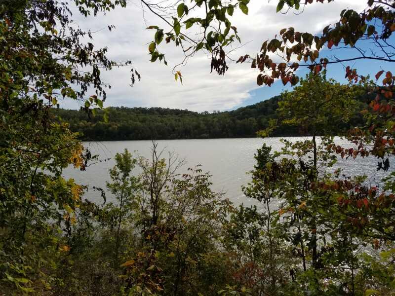 Serene view of a lake surrounded by lush greenery and trees, with distant hills and a partly cloudy sky. Dillon State Park mountain bike trail.