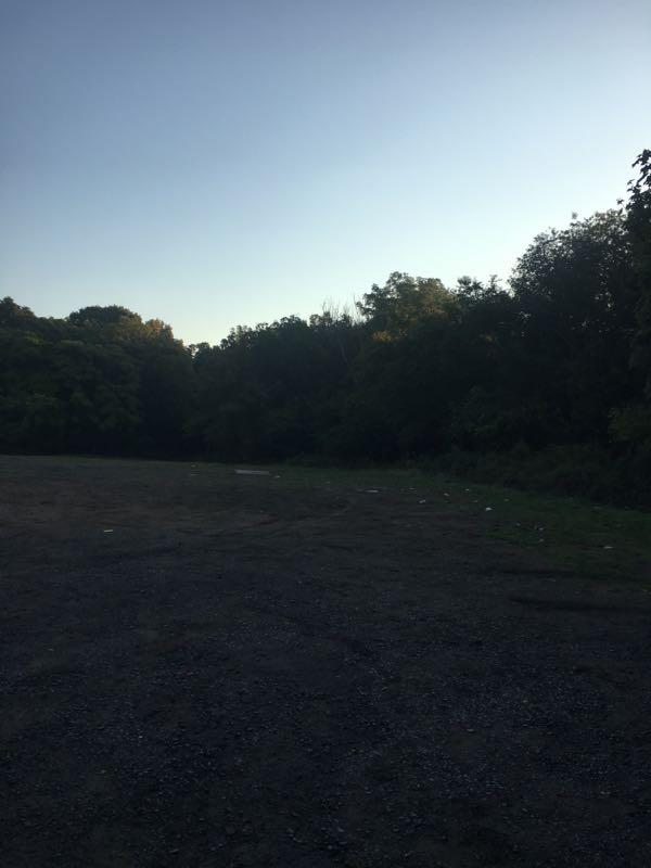 An empty outdoor area with a gravel surface, bordered by dense trees on one side. The sky is clear, transitioning from daylight to dusk. Richmond Avenue and Forest Hill road mountain bike trail.