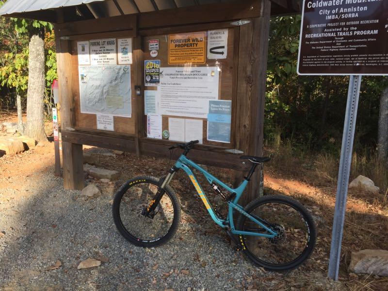 A mountain bike stands parked on gravel in front of an information board in a wooded area. The board displays various notices and maps related to local trails and recreational activities. Signs indicate that the area is part of the Coldwater Mountain project, with warnings and guidelines for visitors. Lush greenery surrounds the scene, creating a natural outdoor setting. Coldwater Mountain mountain bike trail.