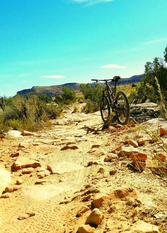 A mountain bike parked on a rugged dirt trail surrounded by sparse vegetation and rocky terrain, with a clear blue sky and distant hills in the background. Klondike Bluffs mountain bike trail.