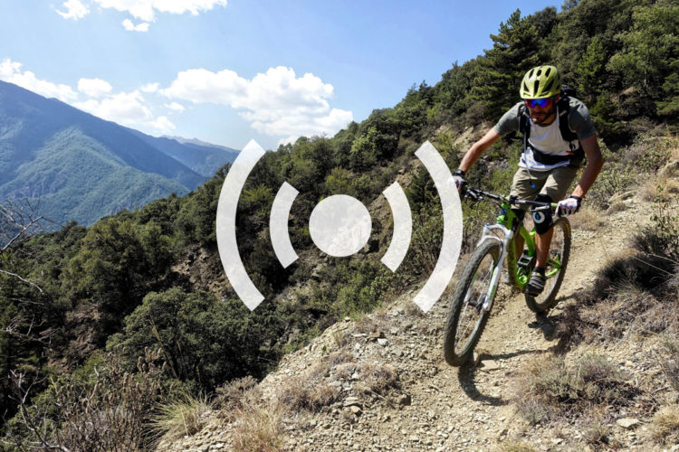A mountain biker navigates a rocky trail surrounded by lush greenery and mountains in the background, under a partially cloudy blue sky.