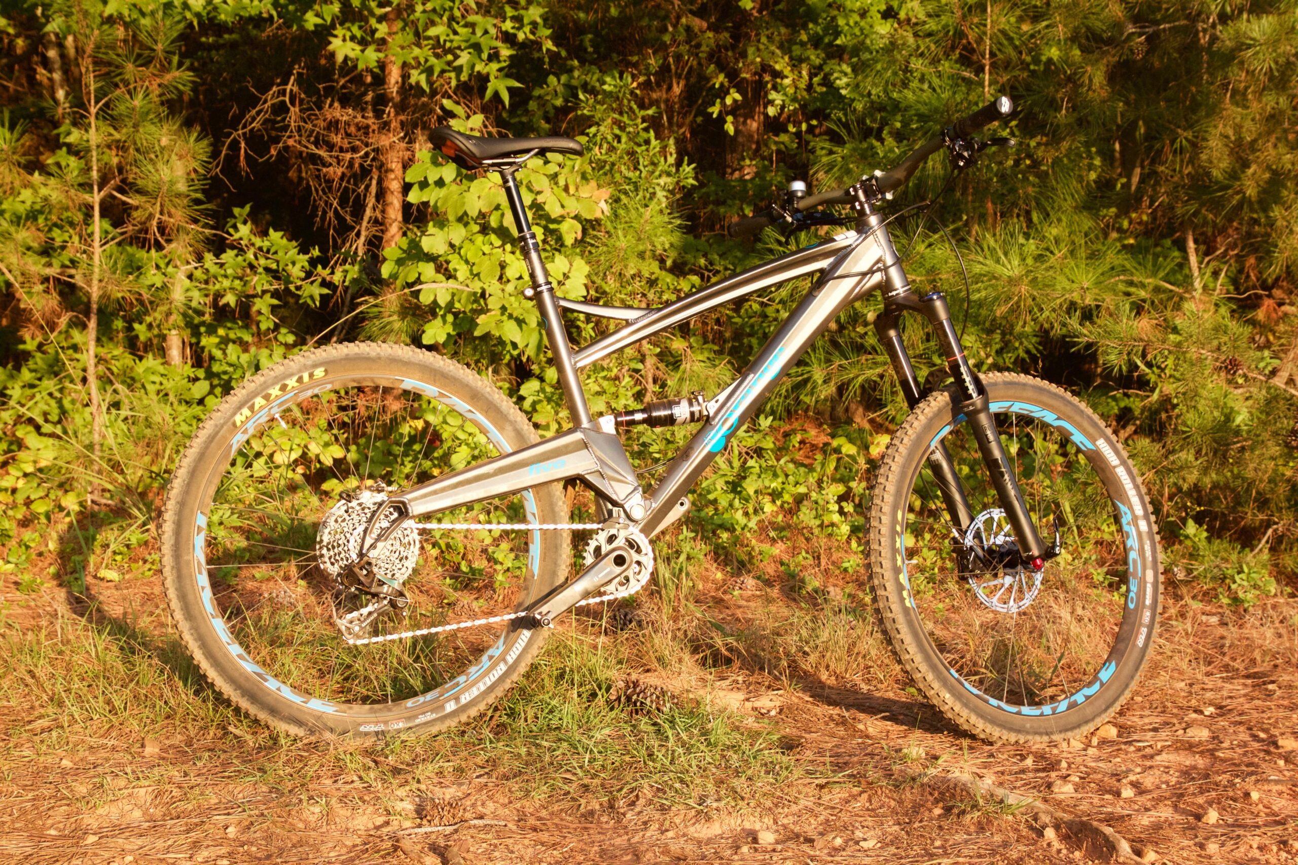 Orange Five RS: A silver mountain bike with blue accents, positioned on a dirt path surrounded by greenery. The bike features a dual suspension system, a prominent gear system on the rear wheel, and knobby tires suitable for off-road riding. Sunlight casts a warm glow on the scene, highlighting the bike's sleek design.