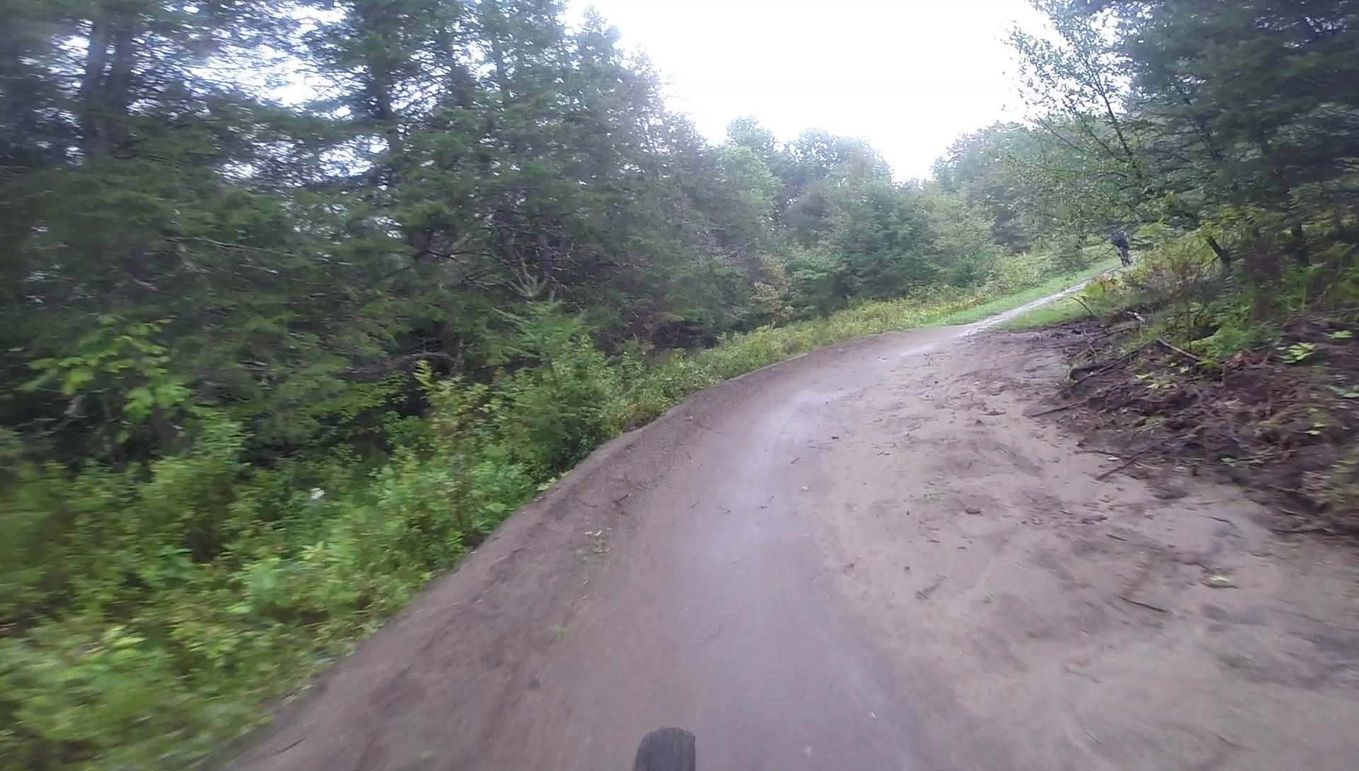 A winding dirt trail surrounded by lush greenery and trees, showing a path that curves to the right. The scene is slightly overcast and appears to be in a forested area, indicating a peaceful outdoor setting for biking or hiking. Kingdom Trails mountain bike trail.