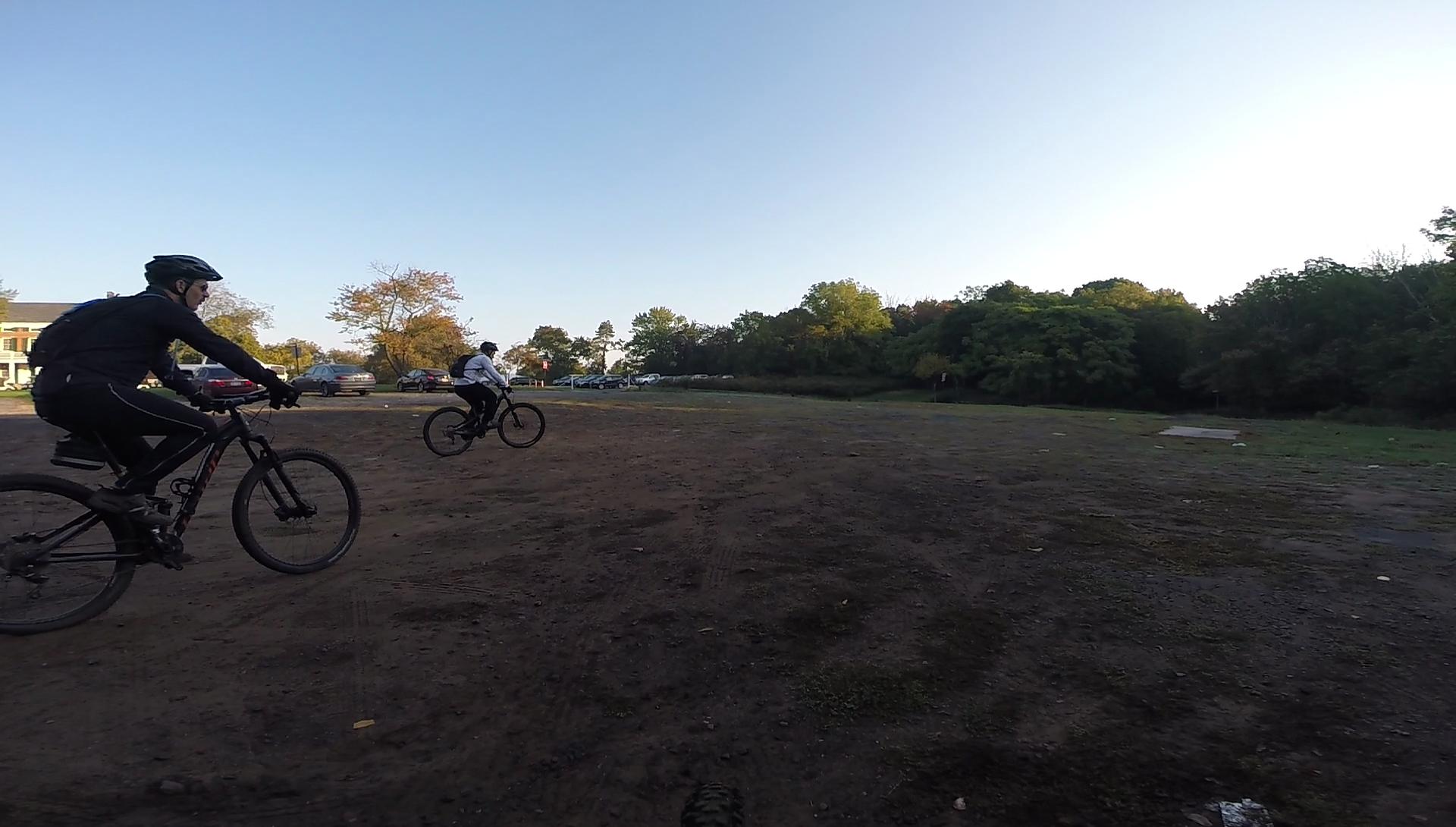 Two mountain bikers riding on a dirt path in a park during the day. The landscape features trees and a clear blue sky, with a parking area visible in the background. One biker is wearing a black jacket and helmet, while the other is in a white jacket. Richmond Avenue and Forest Hill road mountain bike trail.
