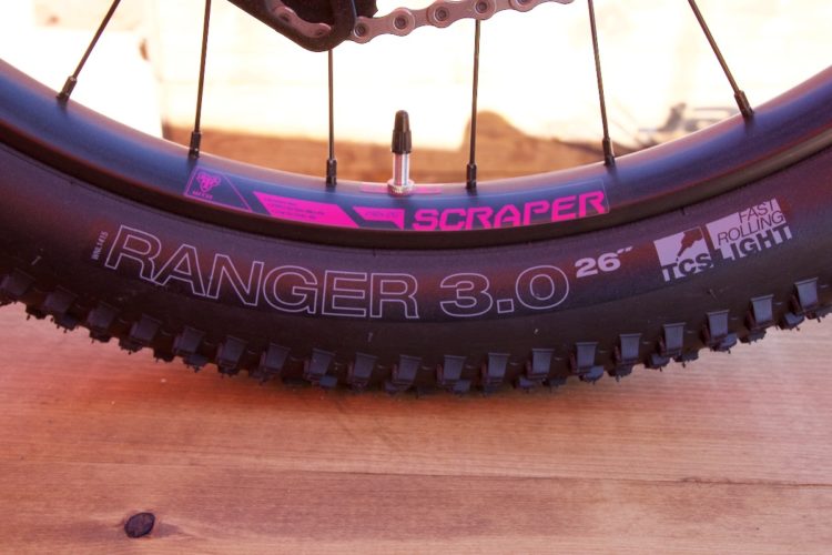 Image of a bicycle tire featuring the brand name "SCRAPER" in pink at the top and "RANGER 3.0 26”" prominently displayed in white on the sidewall. The tire has a knobby design suitable for off-road biking, and it is set against a wooden surface.