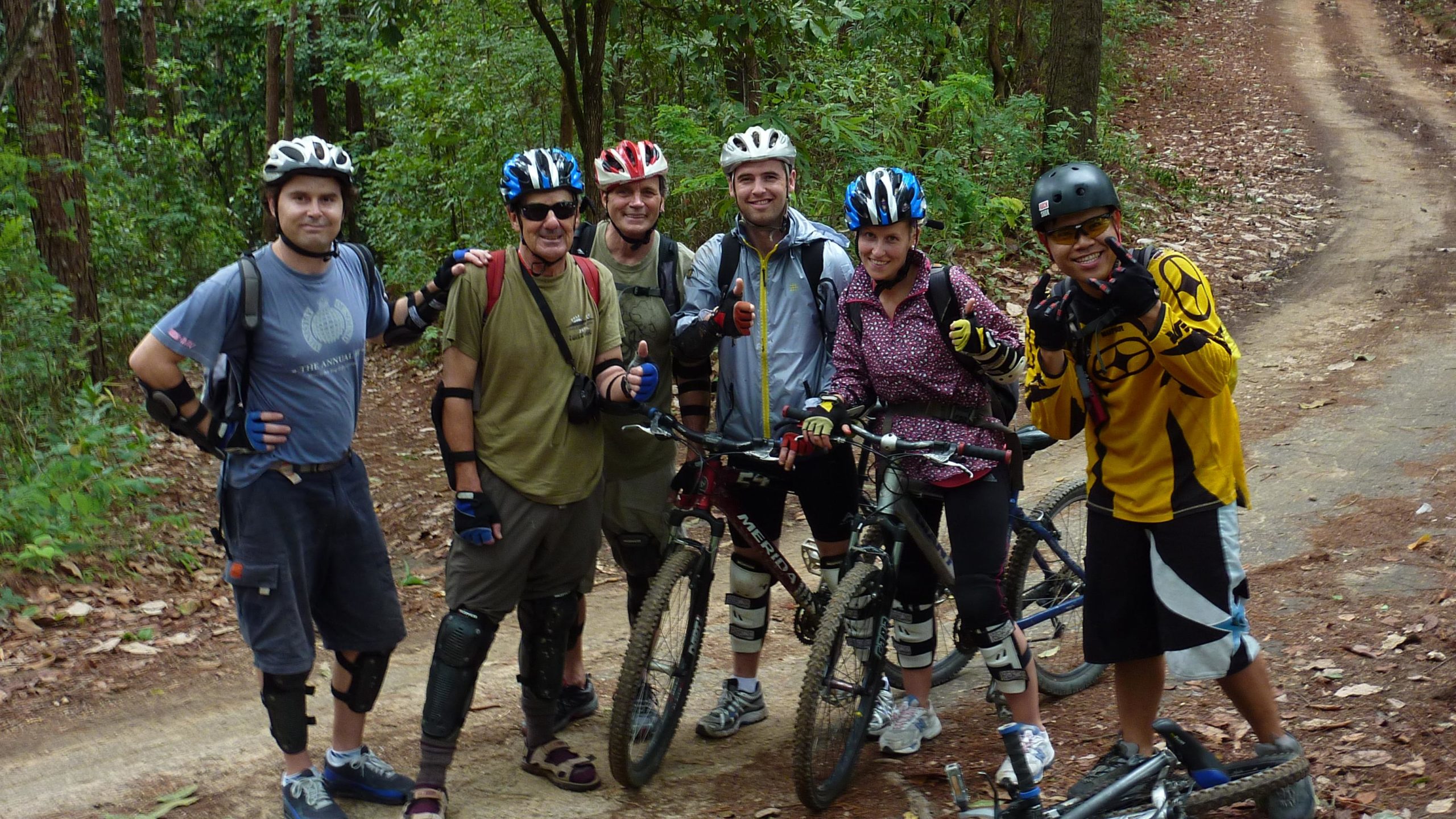 A group of five people, wearing helmets and protective gear, pose for a photo on a wooded mountain biking trail. They are standing with their bikes, smiling and giving thumbs up. The surrounding area features lush green foliage, and the dirt path winds through the trees. The Eagles Route mountain bike trail.