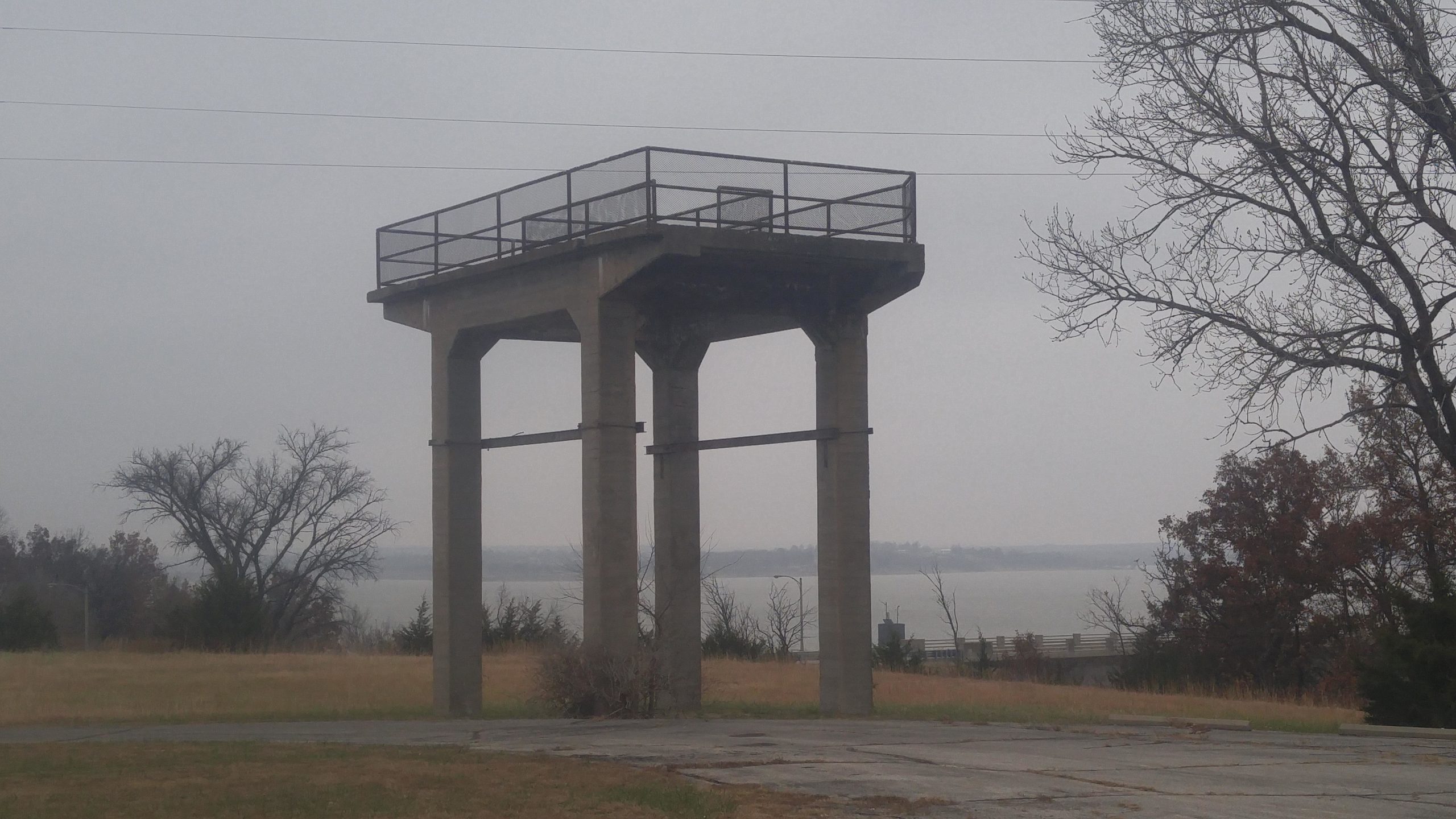 A concrete observation tower stands on a grassy area by a lake, surrounded by sparse trees. The sky is overcast, giving the scene a gray, subdued ambiance. The lake is partially visible in the background. Fall River State Park mountain bike trail.