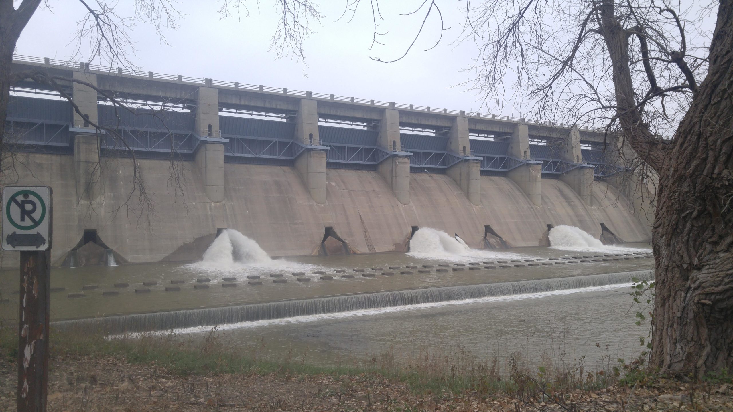 A concrete dam with water flowing through several release gates, surrounded by a river. The top section has a blue structural overlay and a walkway, while the foreground shows a grassy area and a parking sign. The sky appears overcast, creating a gray atmosphere. Fall River State Park mountain bike trail.
