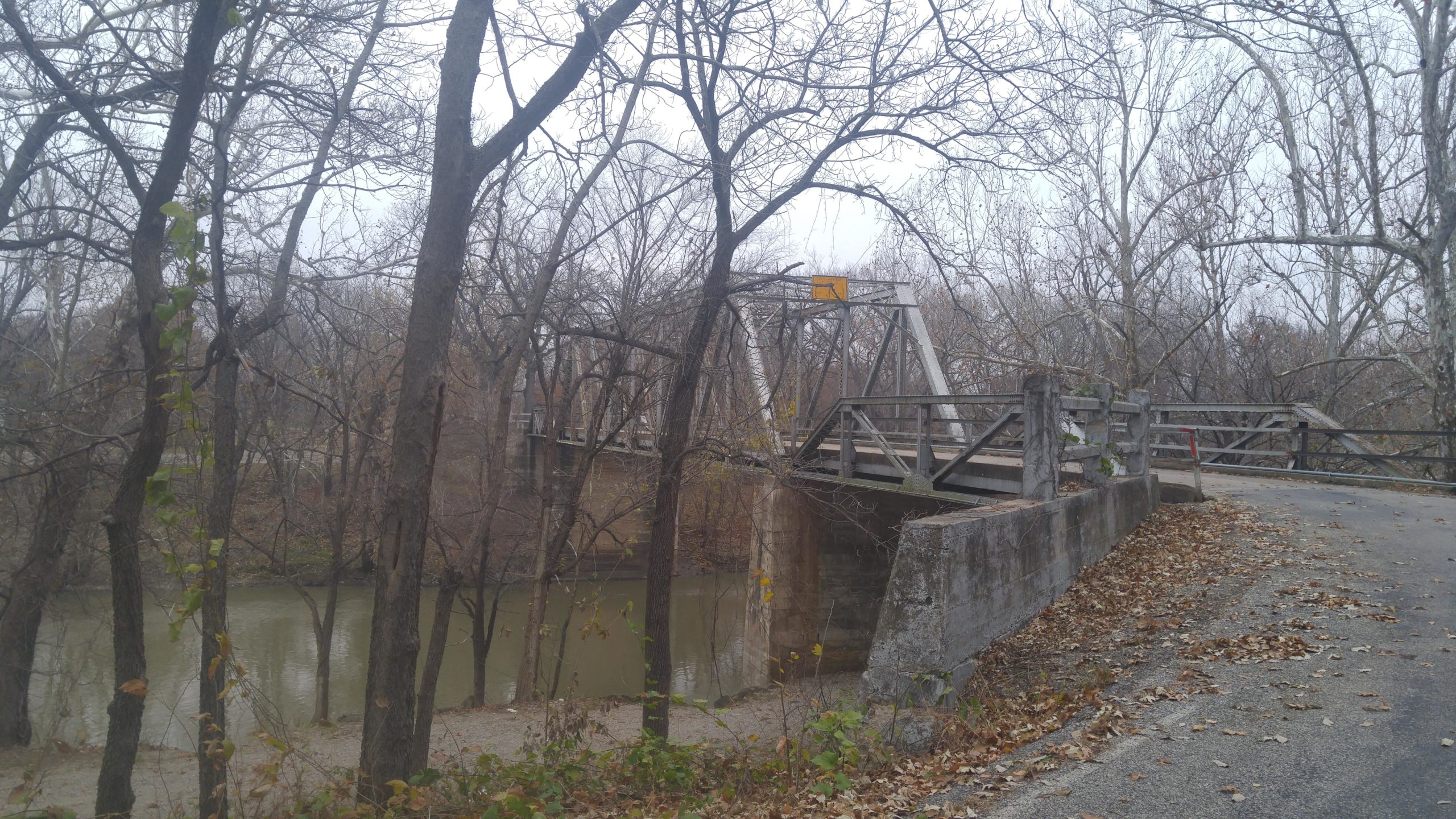 A weathered metal bridge spans a calm, greenish river, surrounded by bare trees in a rural setting. The scene is slightly overcast, with fallen leaves scattered along a gravel path leading up to the bridge. The structure features an overhead warning sign and has a rustic, aged appearance. Fall River State Park mountain bike trail.