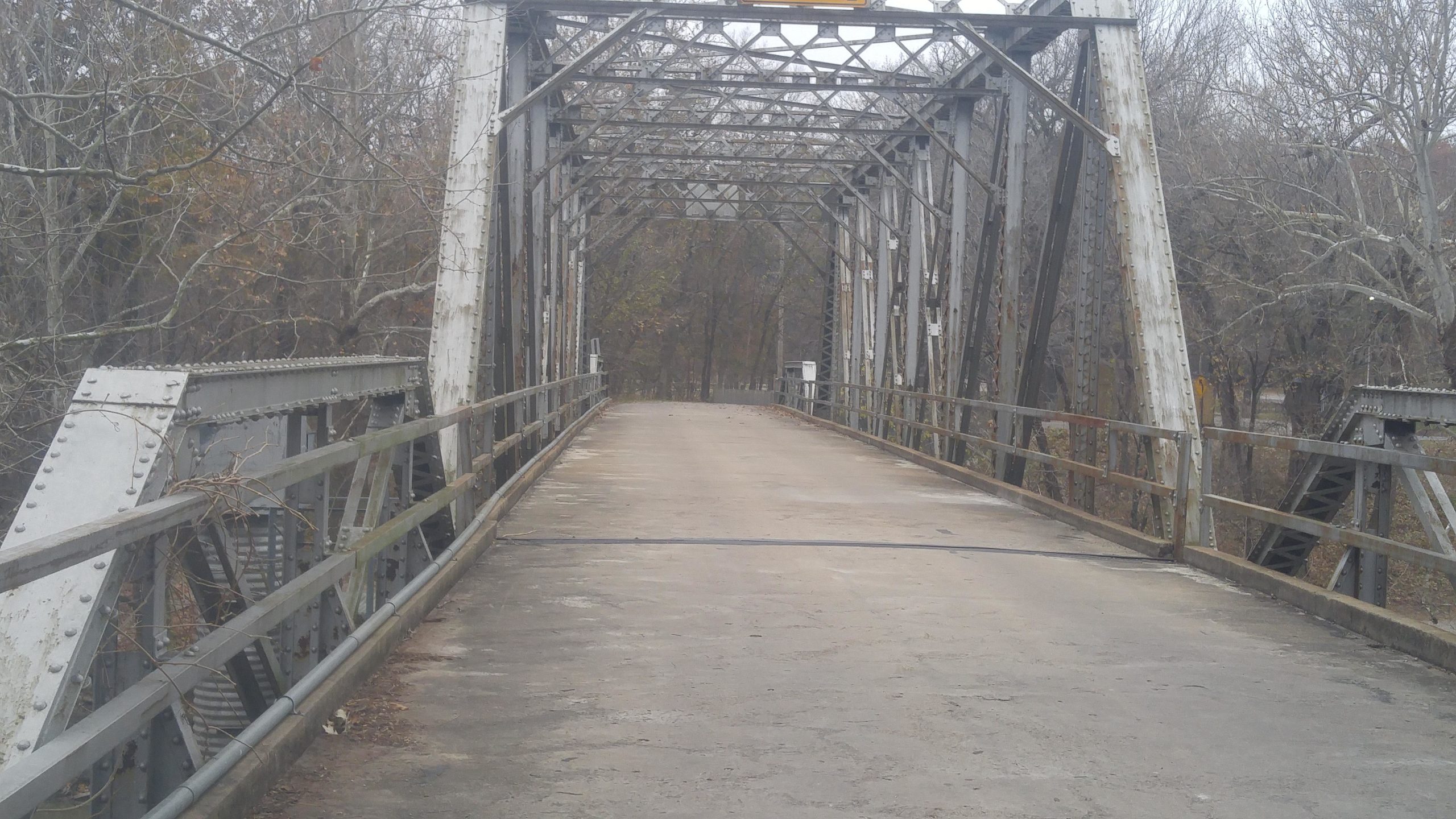 Alt text: A weathered, steel truss bridge with a concrete surface, surrounded by bare trees. The bridge has a handrail along the sides and is empty, with no vehicles or pedestrians. The sky is overcast, adding a somber mood to the scene. Fall River State Park mountain bike trail.