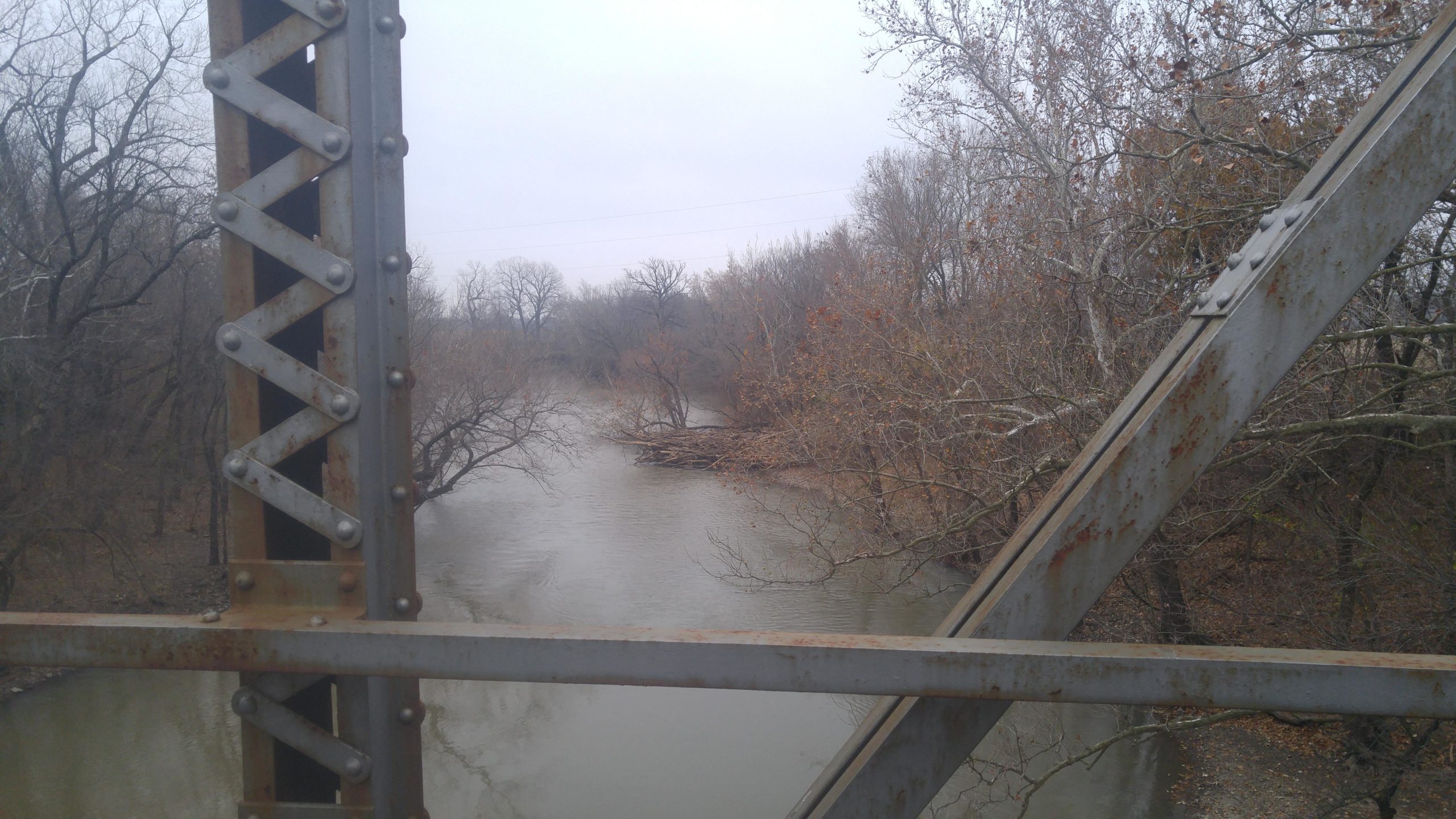 Alt text: A view of a river from an elevated position on a rusty metal bridge, with bare trees lining the banks and a cloudy sky in the background. The water is calm, and some fallen branches are visible along the shore. Fall River State Park mountain bike trail.