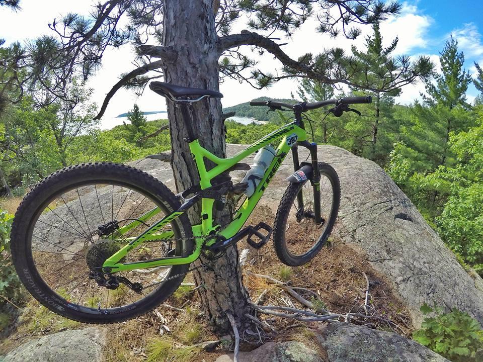 Trek trek remedy 7 27.5: A green mountain bike leaning against a tree on a rocky outcrop, surrounded by lush greenery and distant hills under a partly cloudy sky.