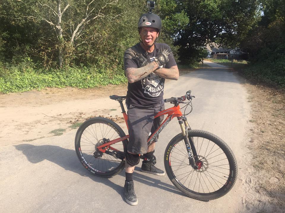 Santa Cruz Tallboy Carbon: A person wearing a helmet and a graphic t-shirt stands next to a mountain bike on a dirt path, playfully sticking out their tongue and making a hand gesture. The background features greenery and trees, suggesting an outdoor environment.