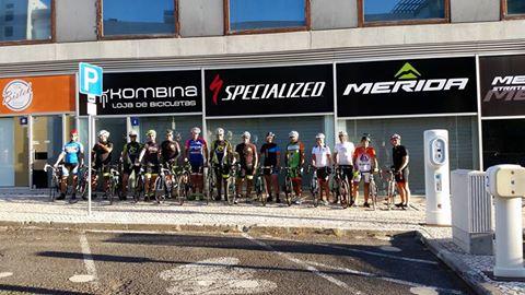 A group of cyclists posing with their bikes in front of a bike shop, featuring logos of various brands including Specialized and Merida. The scene is set on a sunny day, with a line of parked bicycles and shop signage prominently displayed.