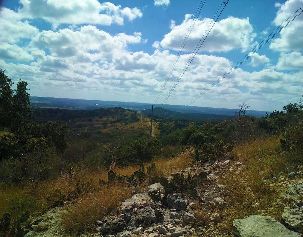 A scenic landscape featuring rolling hills under a partly cloudy sky. In the foreground, there are rocky outcrops and cacti, while power lines stretch across the midground leading towards the horizon. The background showcases a vast expanse of greenery and open fields, creating a tranquil natural setting. Flat Rock Ranch mountain bike trail.