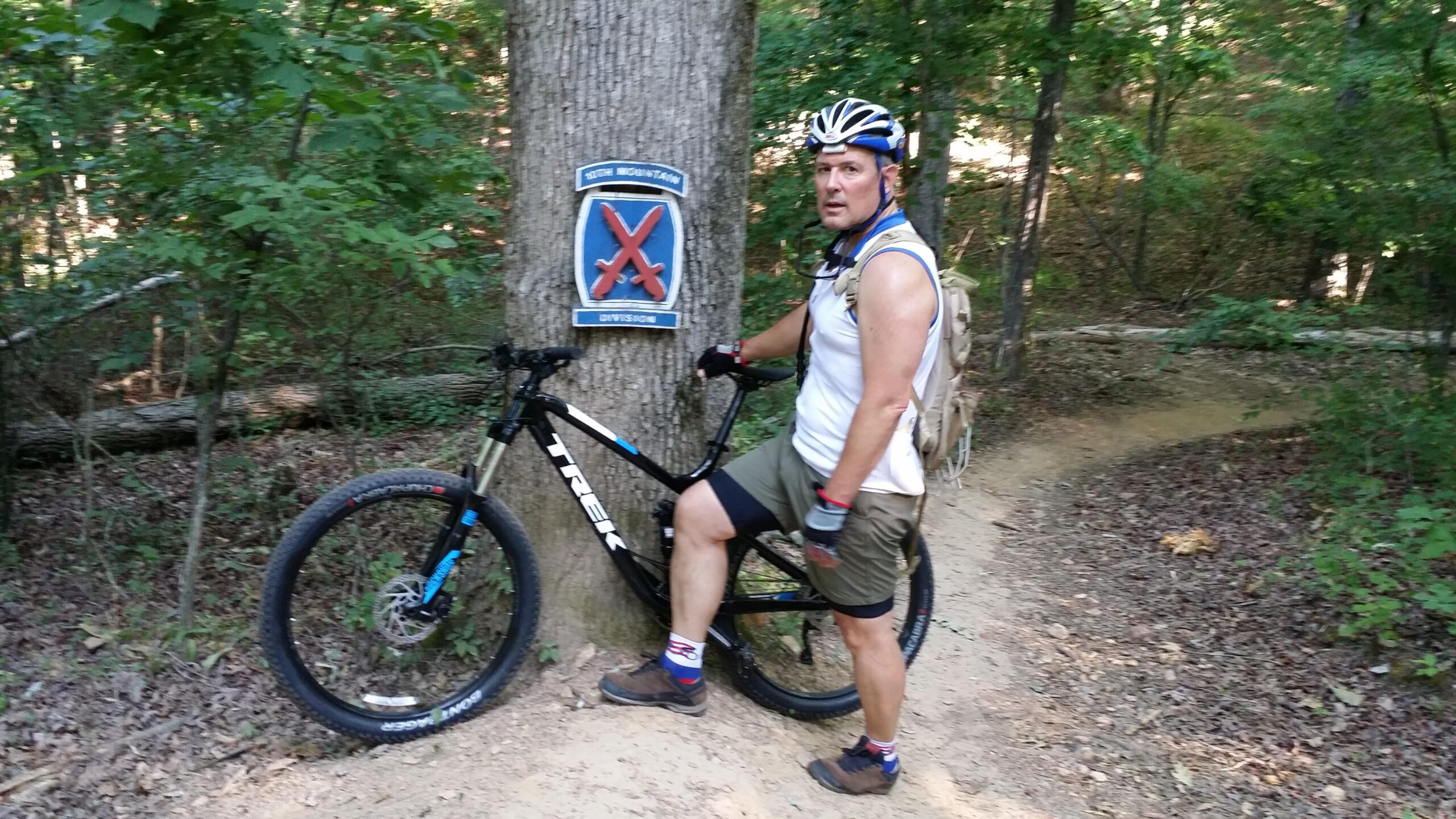 Trek Fuel EX: A person in athletic clothing, wearing a helmet and gloves, stands next to a mountain bike leaned against a tree. The background features a lush wooded area with a dirt path. A sign on the tree indicates the "10th Mountain Division."