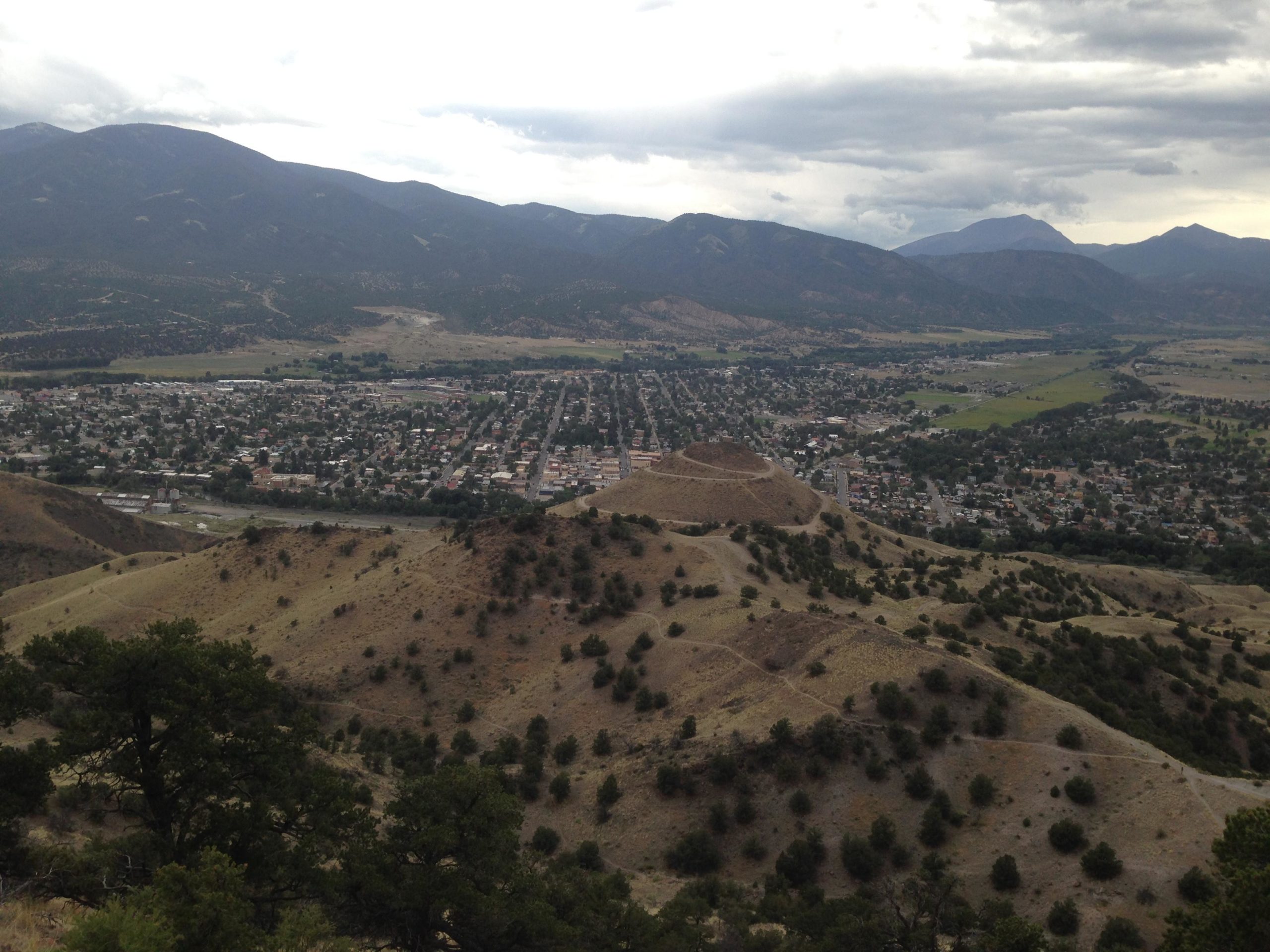 Aerial view of a small town nestled in a valley, surrounded by rolling hills and mountains under a partly cloudy sky. The town features a grid of streets and residential buildings, with patches of greenery visible throughout. The landscape includes dry, grassy hills and scattered trees, creating a natural contrast to the urban area below. Arkansas Hills mountain bike trail.