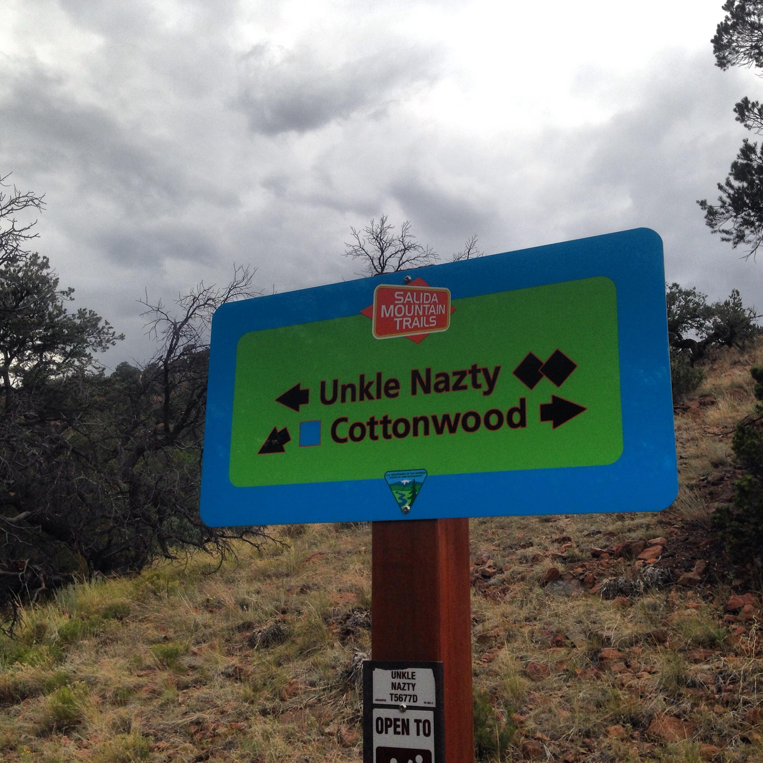 Signpost for Salida Mountain Trails, indicating the directions to Unkle Nazty and Cottonwood trails, with icons representing trail difficulty. The background features a cloudy sky and sparse vegetation. Arkansas Hills mountain bike trail.