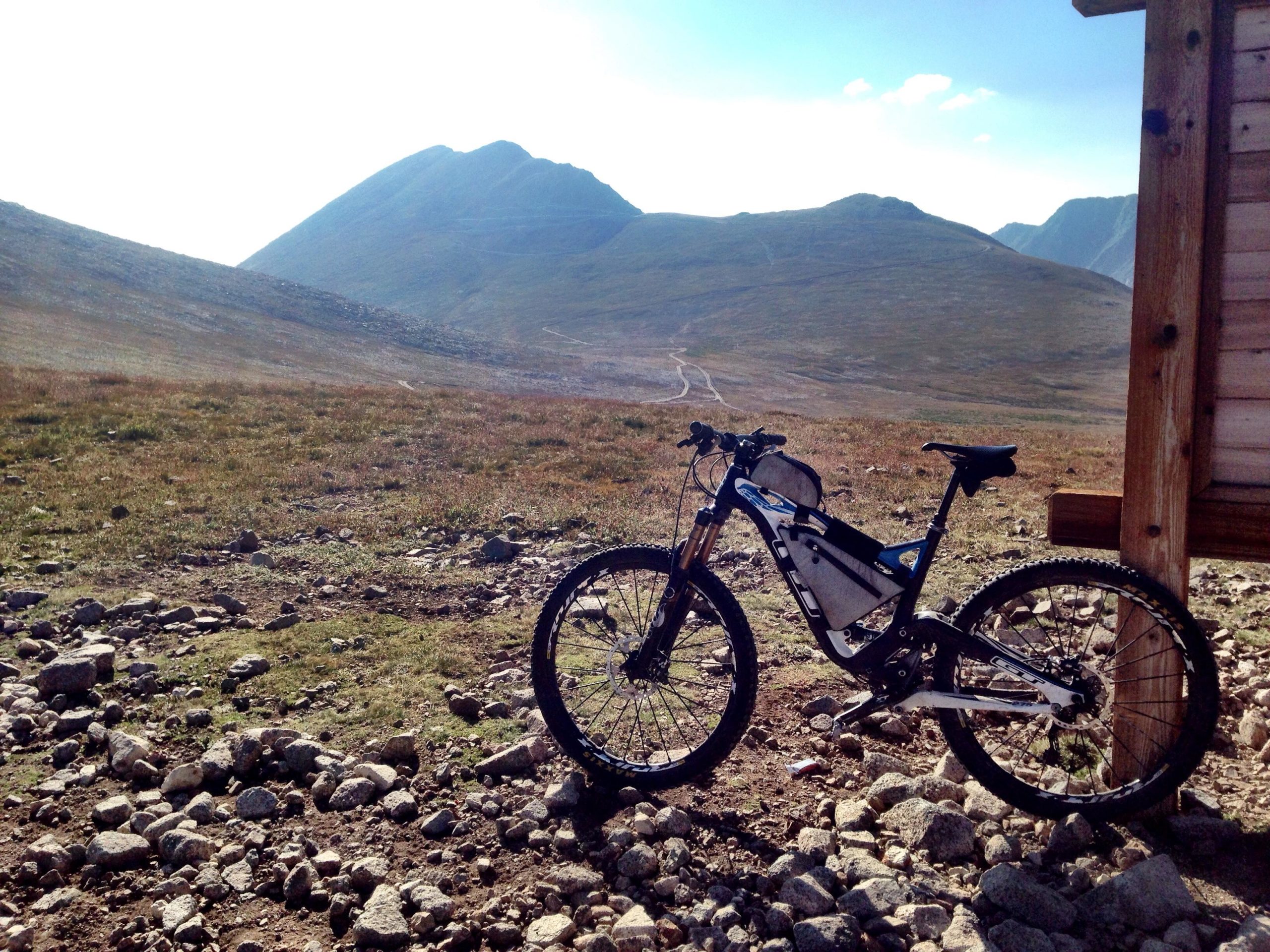 A mountain bike resting on rocky terrain with a mountainous landscape in the background. The bike is positioned near a wooden structure, and the scene features a clear sky and rolling hills, suggesting a remote outdoor location. Mount White Road / FS #278.B mountain bike trail.