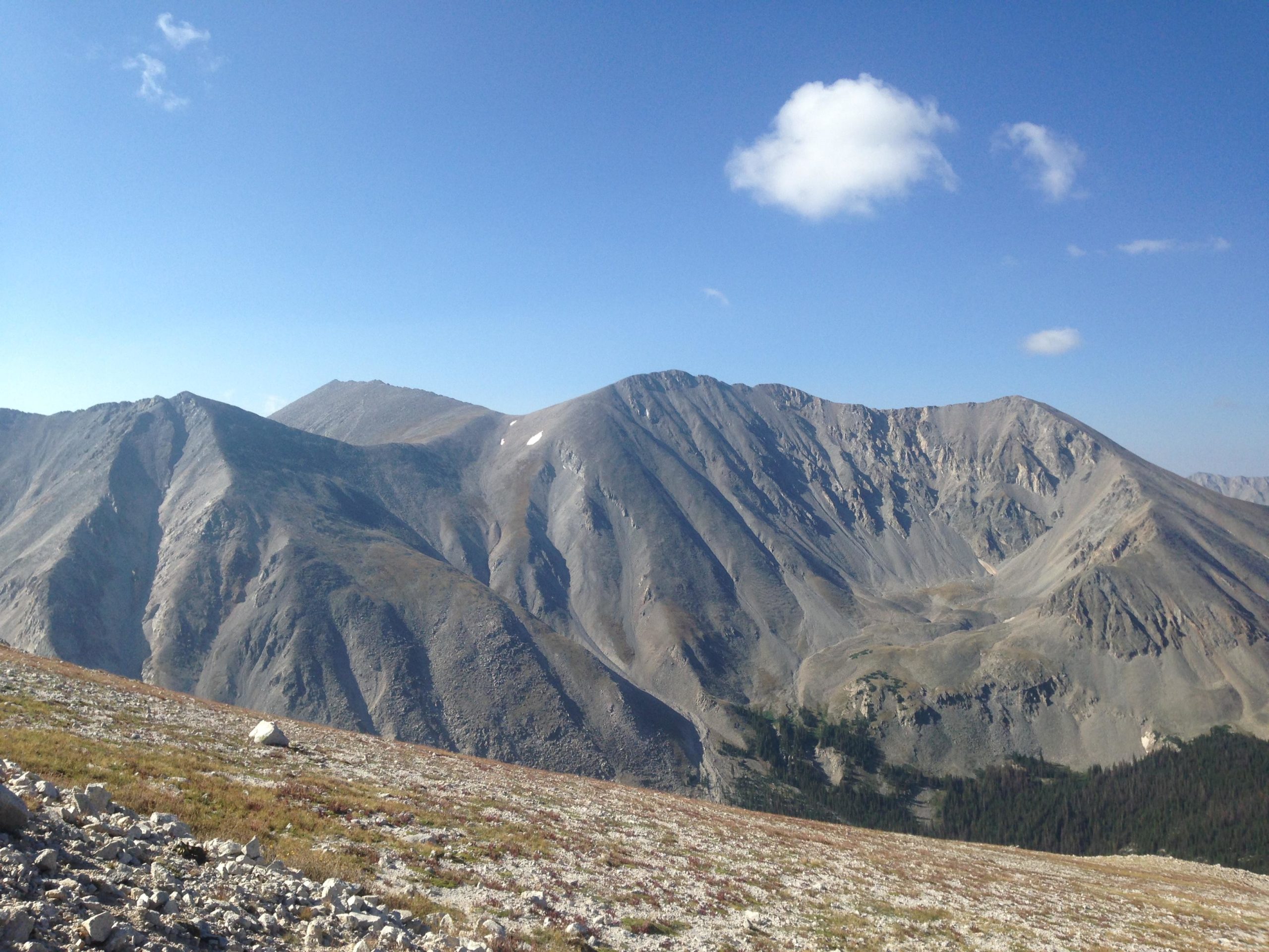 A panoramic view of rugged mountain ranges under a clear blue sky, with gently rolling slopes and patches of greenery visible in the valleys. The landscape displays a mix of rocky terrain and sparse vegetation, highlighting the natural beauty of the wilderness. Mount White Road / FS #278.B mountain bike trail.
