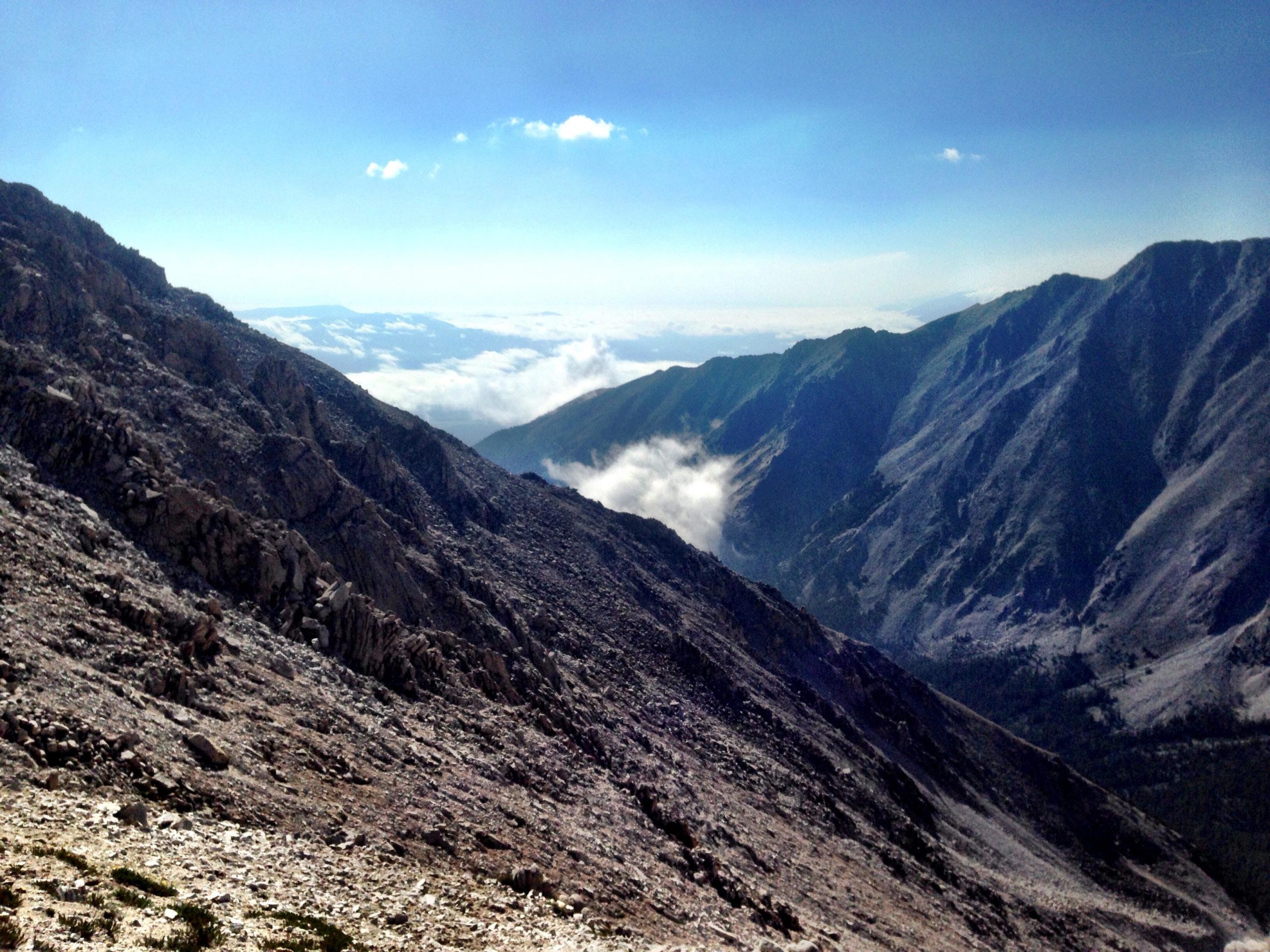 A scenic view of rugged mountain slopes under a clear blue sky, with valleys and clouds visible in the distance. The rocky terrain features steep inclines and varying textures, suggesting a high-altitude environment. Mount White Road / FS #278.B mountain bike trail.