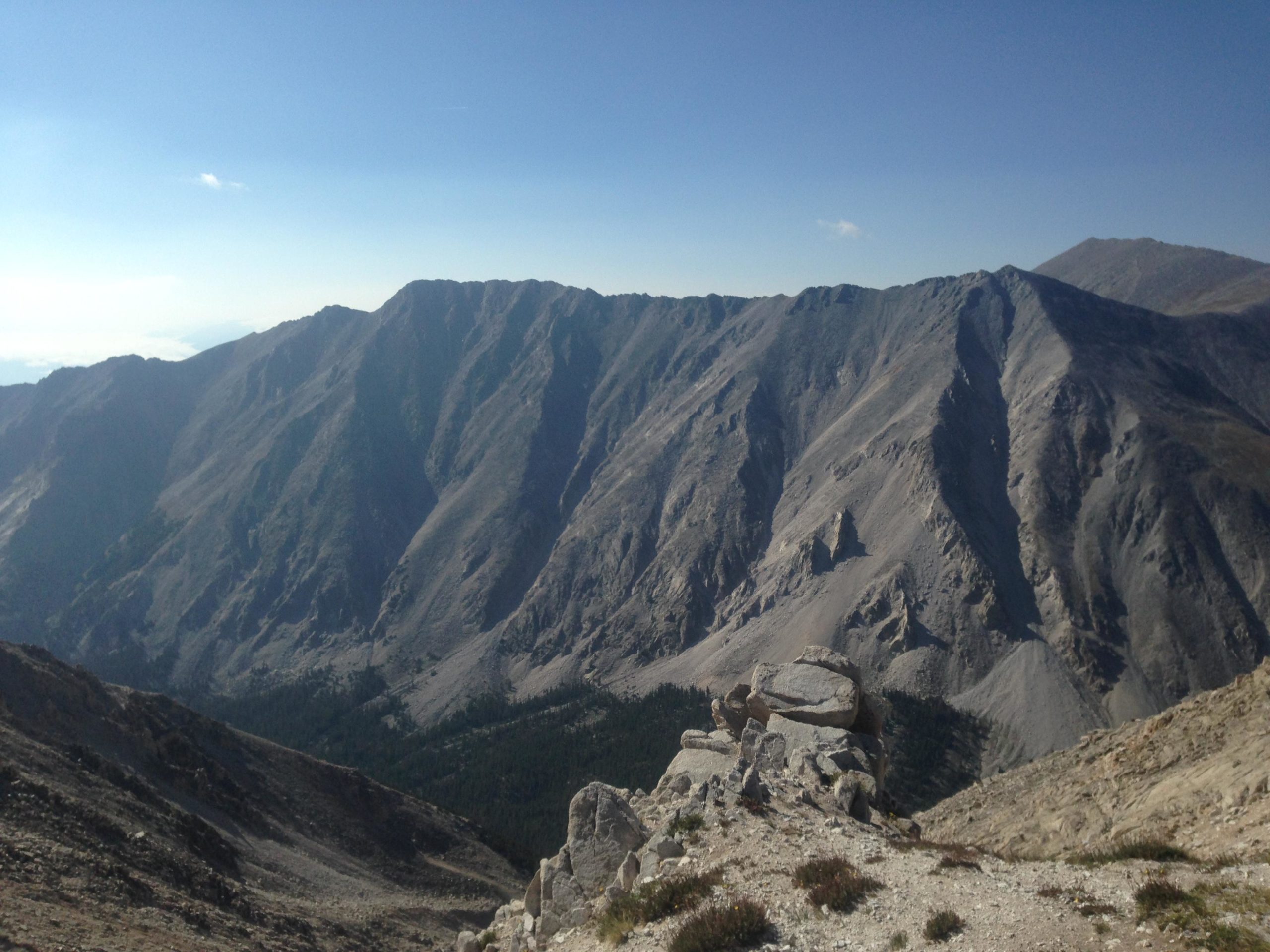 A panoramic view of rugged mountains under a clear blue sky. The landscape features steep, rocky slopes with varying shades of gray and hints of green vegetation in the valleys below. Boulders are visible in the foreground, highlighting the rocky terrain. Mount White Road / FS #278.B mountain bike trail.
