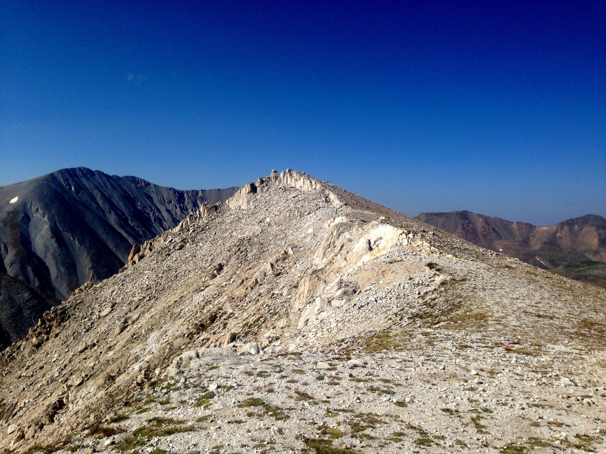 A rocky mountain ridge stretches across the image, with a clear blue sky overhead. The rugged terrain features a mix of loose stones and patches of grass, while distant mountains rise majestically in the background. The overall scene conveys a sense of natural beauty and wilderness. Mount White Road / FS #278.B mountain bike trail.