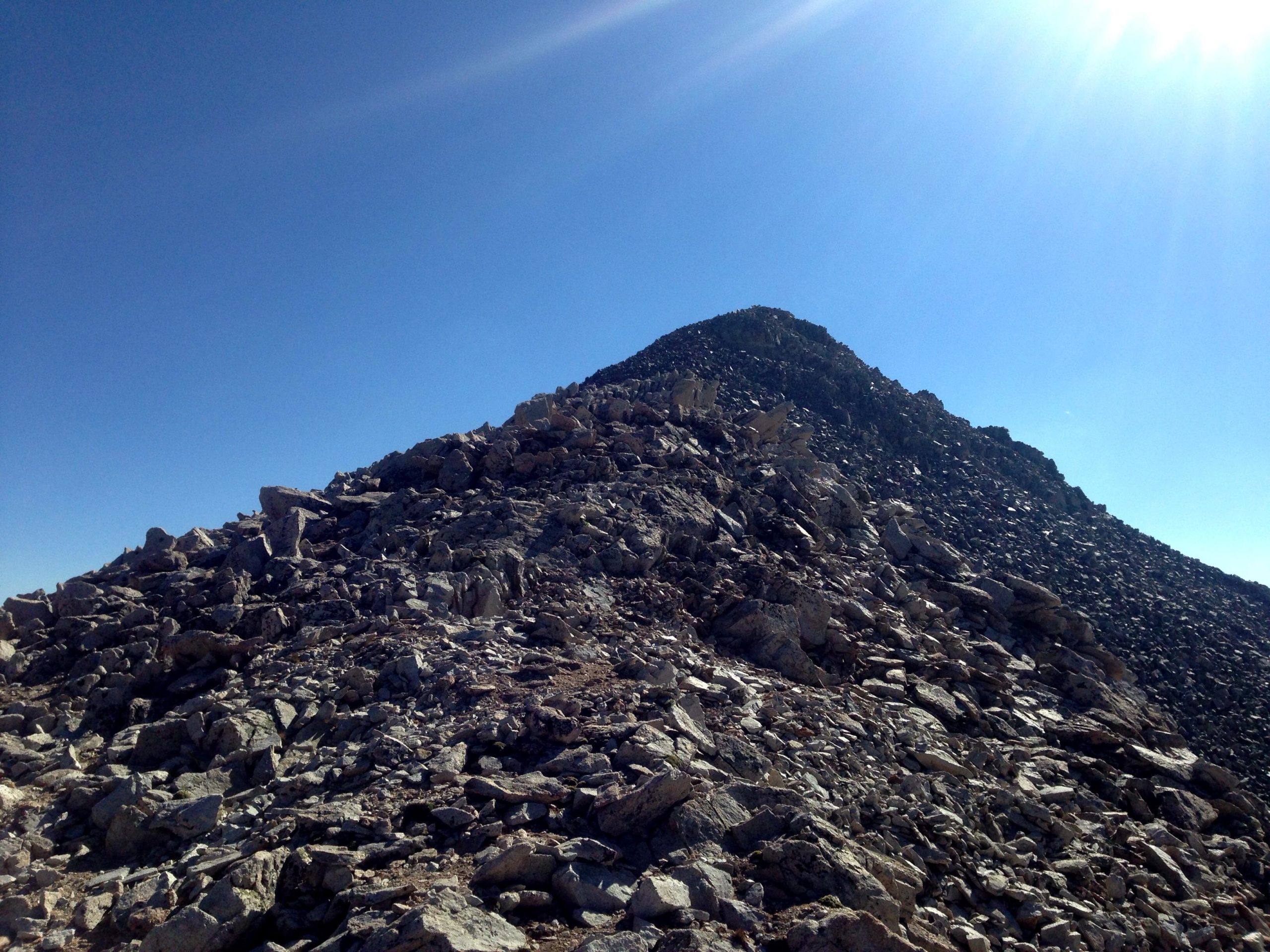 A rocky mountain slope under a clear blue sky, featuring a sharp peak in the distance. The foreground is filled with a variety of large and small boulders, showcasing the rugged terrain. The sun shines brightly, illuminating the landscape. Mount White Road / FS #278.B mountain bike trail.
