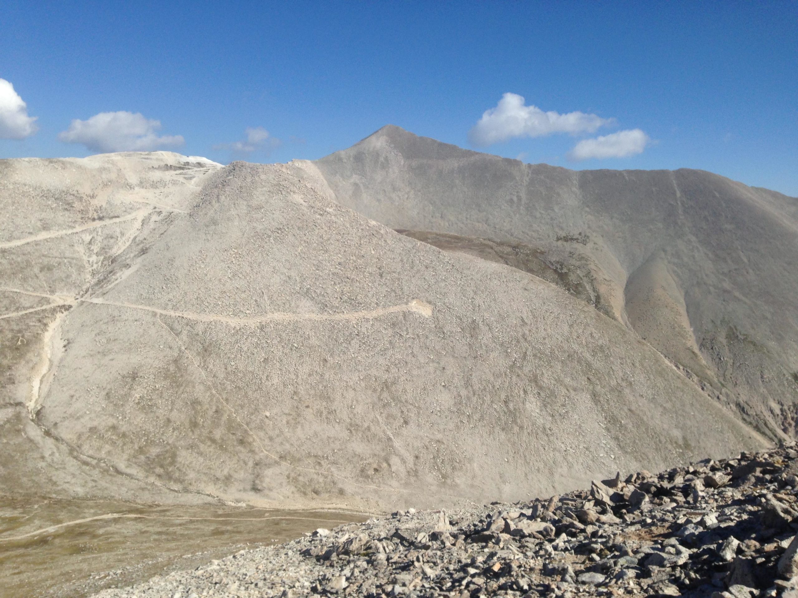 A rugged mountain landscape featuring steep, rocky slopes under a clear blue sky with a few scattered clouds. The terrain is barren with dry, stony ground and visible paths winding through the hills. Mount White Road / FS #278.B mountain bike trail.