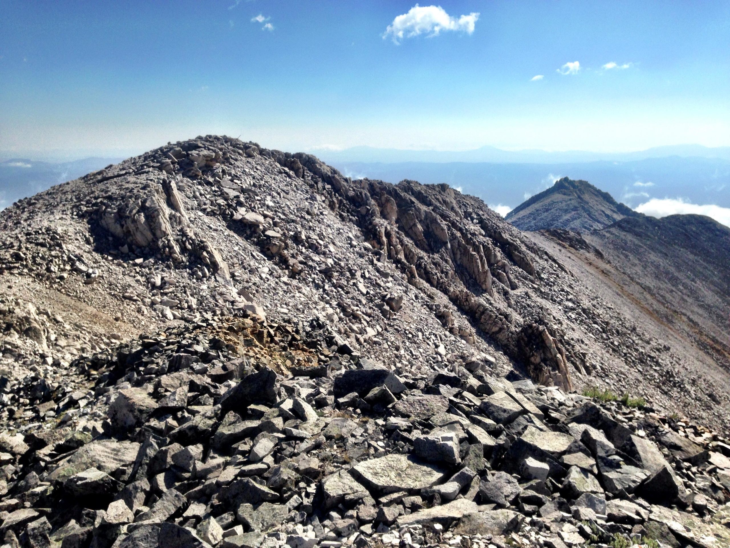 A rocky mountain landscape with multiple peaks under a clear blue sky, showcasing a mix of large stones and gravel on the terrain. The foreground features a rocky outcrop, leading to sloping hills in the background, with distant mountain ranges visible on the horizon. Mount White Road / FS #278.B mountain bike trail.