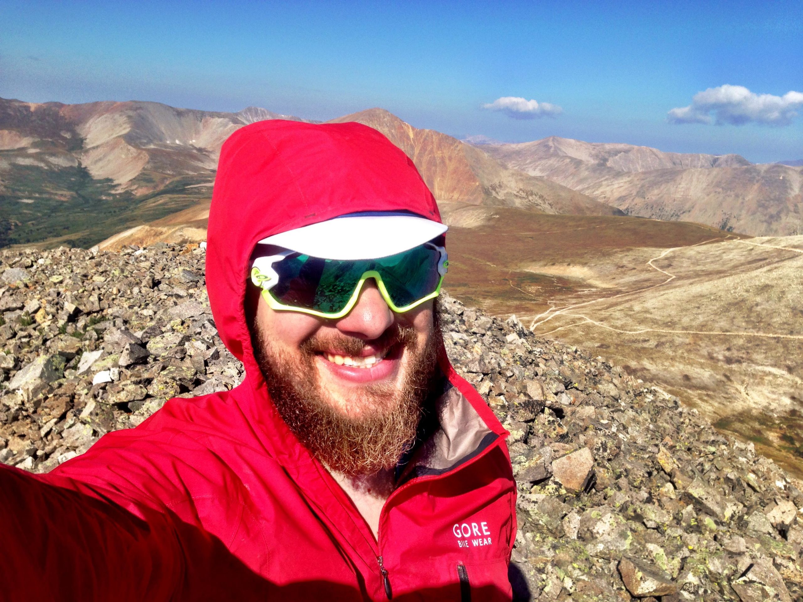 A smiling person wearing a red waterproof jacket and sunglasses stands on a rocky mountain peak, with a scenic landscape of rolling hills and blue skies in the background. Mount White Road / FS #278.B mountain bike trail.