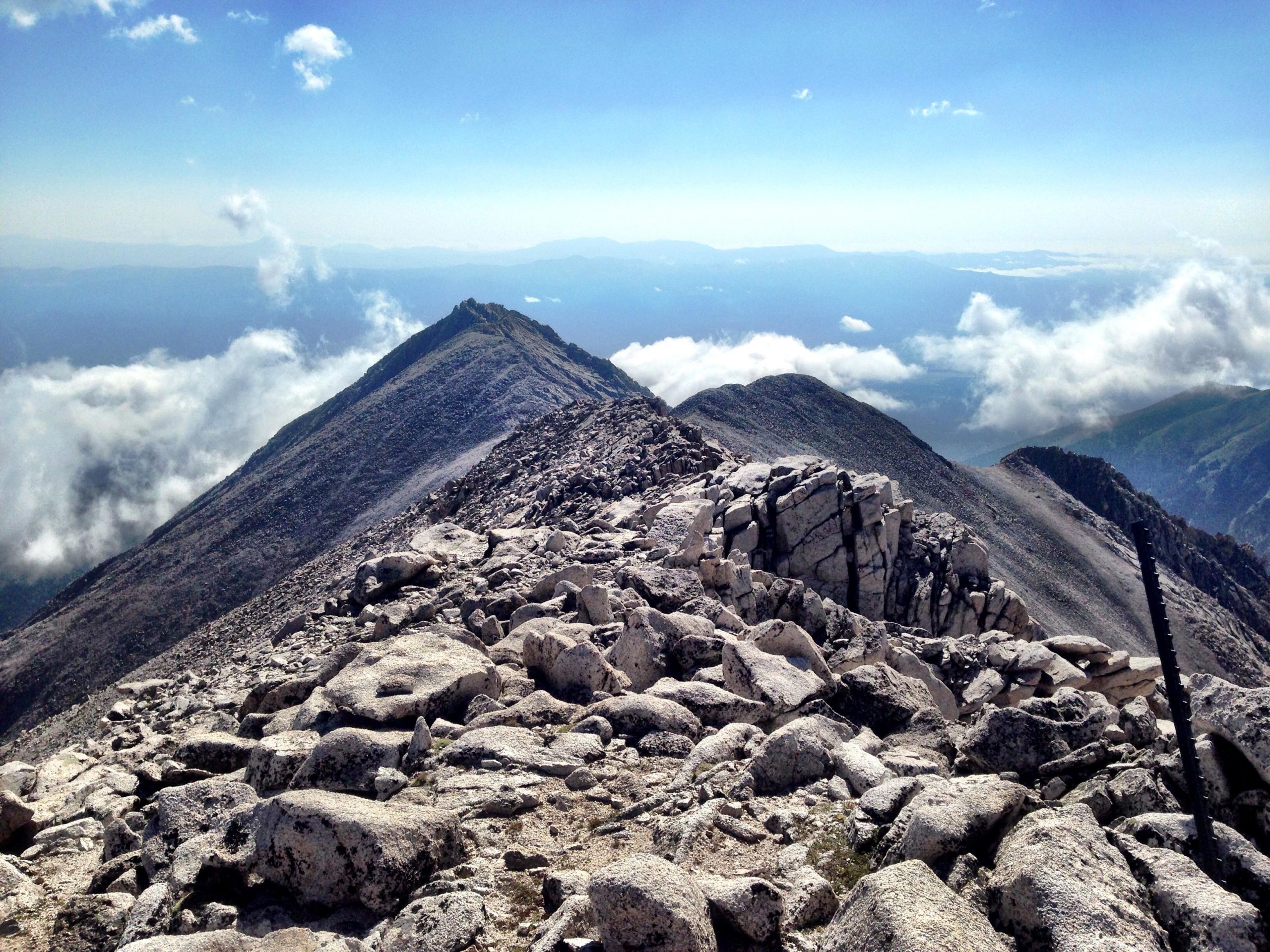 A panoramic view from a mountain peak, showcasing rocky terrain with large boulders in the foreground and steep slopes leading to distant mountainous ridges. Patches of clouds hover below, while the sky is bright with a few scattered clouds. Mount White Road / FS #278.B mountain bike trail.