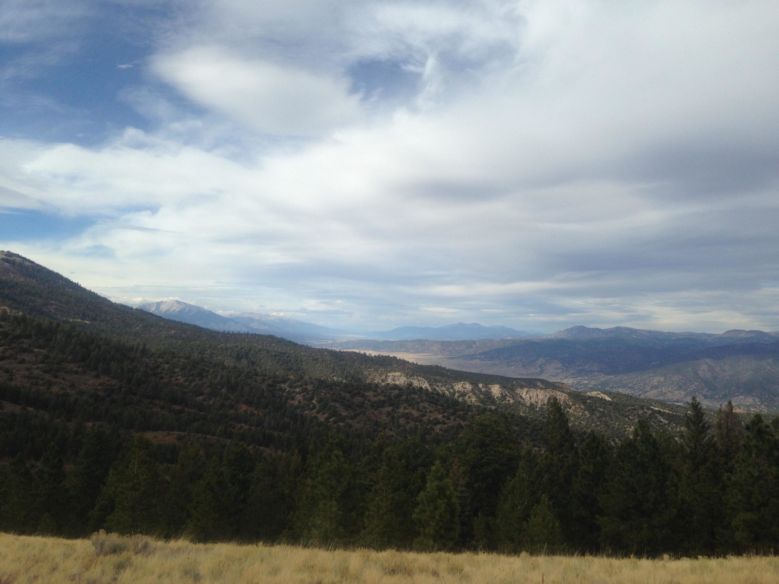 A scenic landscape featuring rolling hills and mountains under a partly cloudy sky. The foreground displays patches of grass and trees, while the mountains in the background are partially covered with clouds. Rainbow Trail: Methodist Mountain Thd to Bear Creek Thd mountain bike trail.
